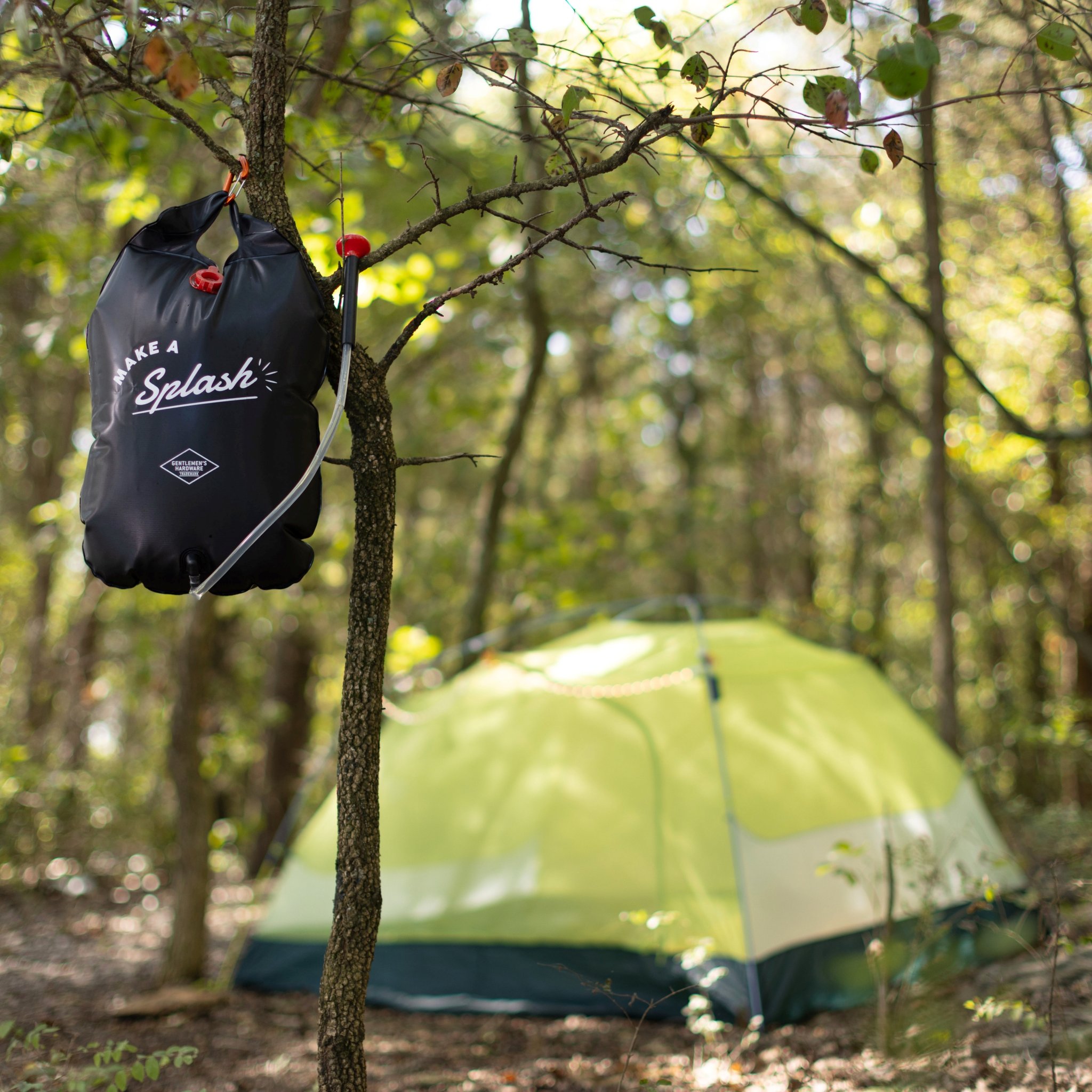 Portable solar shower hung at the left on a tree trunk, a green tent in the middle of a sunny forest clearing
