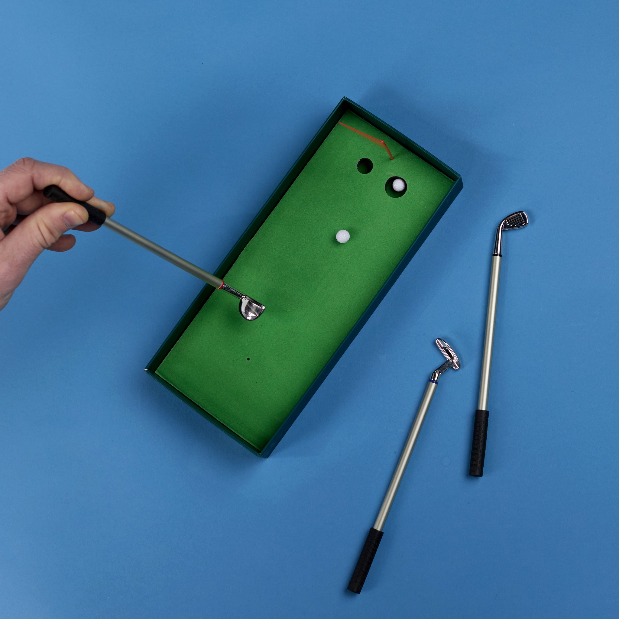 Eine Hand spielt mit einem Golf-Kugelschreiber einen Ball auf dem grünen Mini-Putting-Green der Schachtel