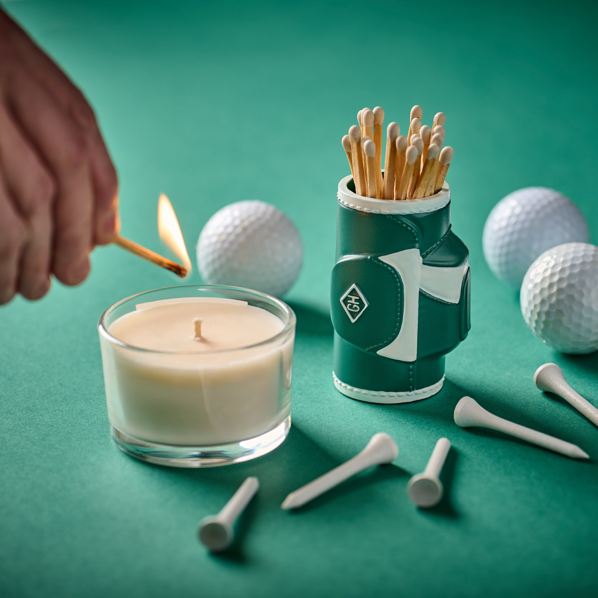 A hand lights a match at a candle in a glass, beside the golf bag match holder, golf balls and tees