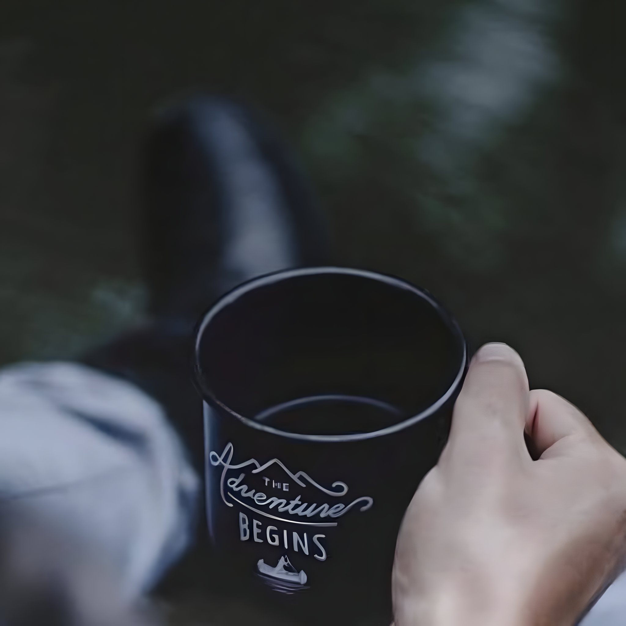Hand hält die Navy Adventure Begins Emailletasse im Wald, im Hintergrund unscharf ein Stiefel und Beine