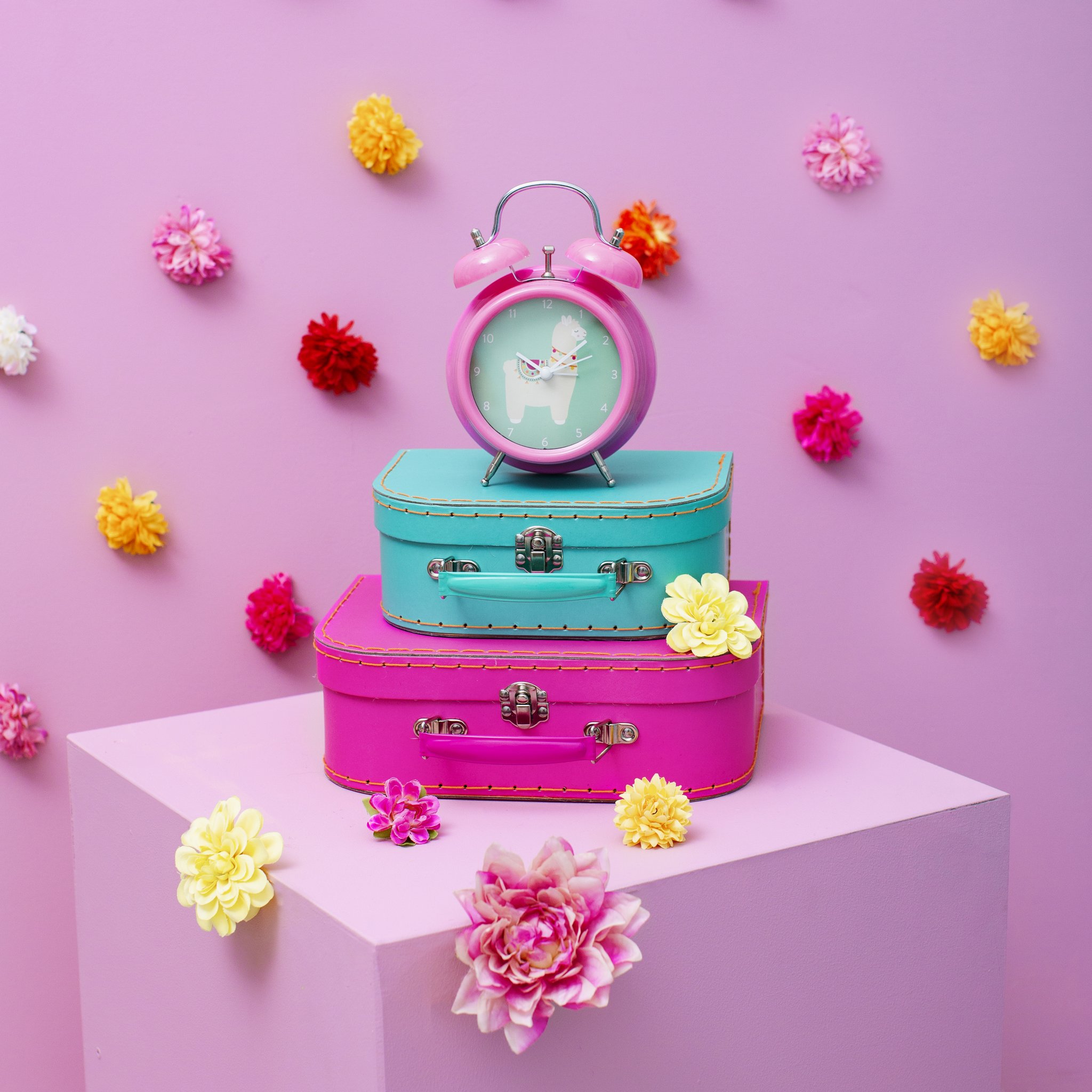 Lima llama alarm clock by Sass & Belle on colourful suitcases with flowers against a pink wall
