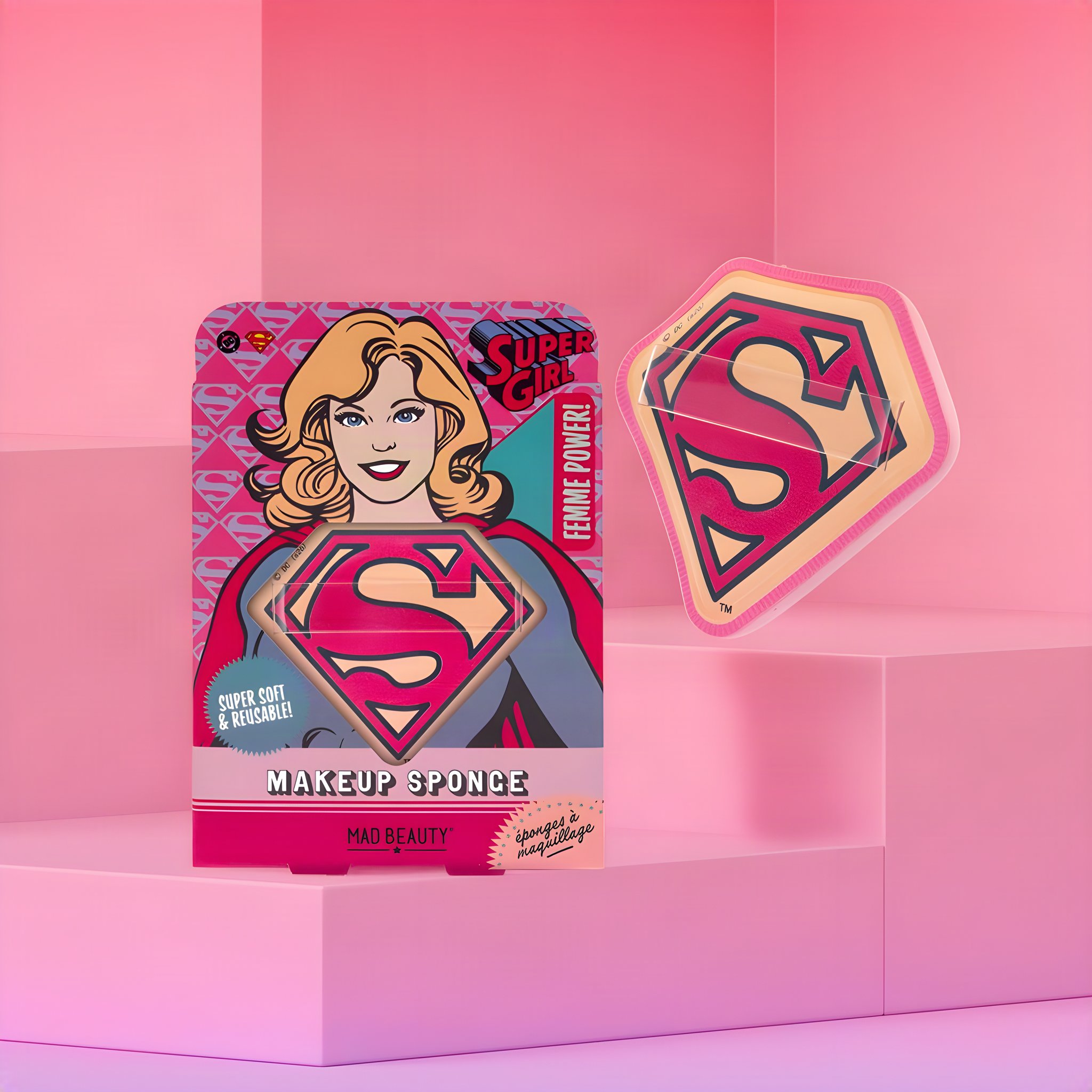 Supergirl Make-up-Schwamm in Schildform mit S-Logo neben der Verpackung mit Comic-Supergirl, rosa Raum