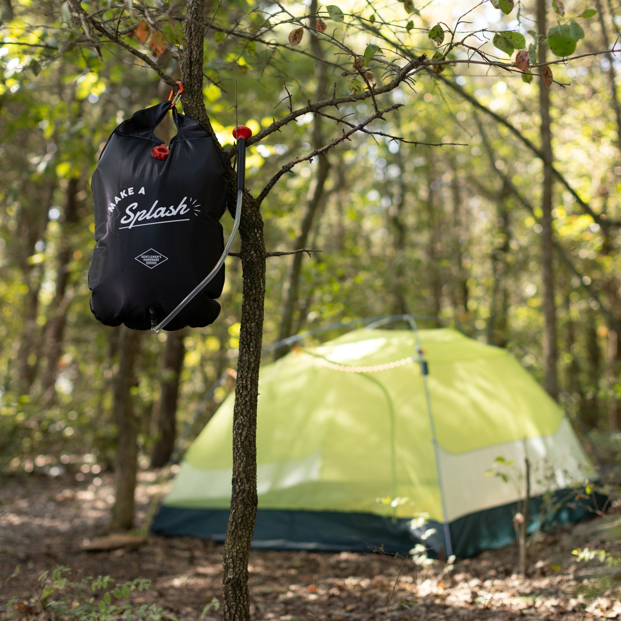 Wider view of the portable solar shower on a tree trunk in a leafy forest, a green tent among the trees below