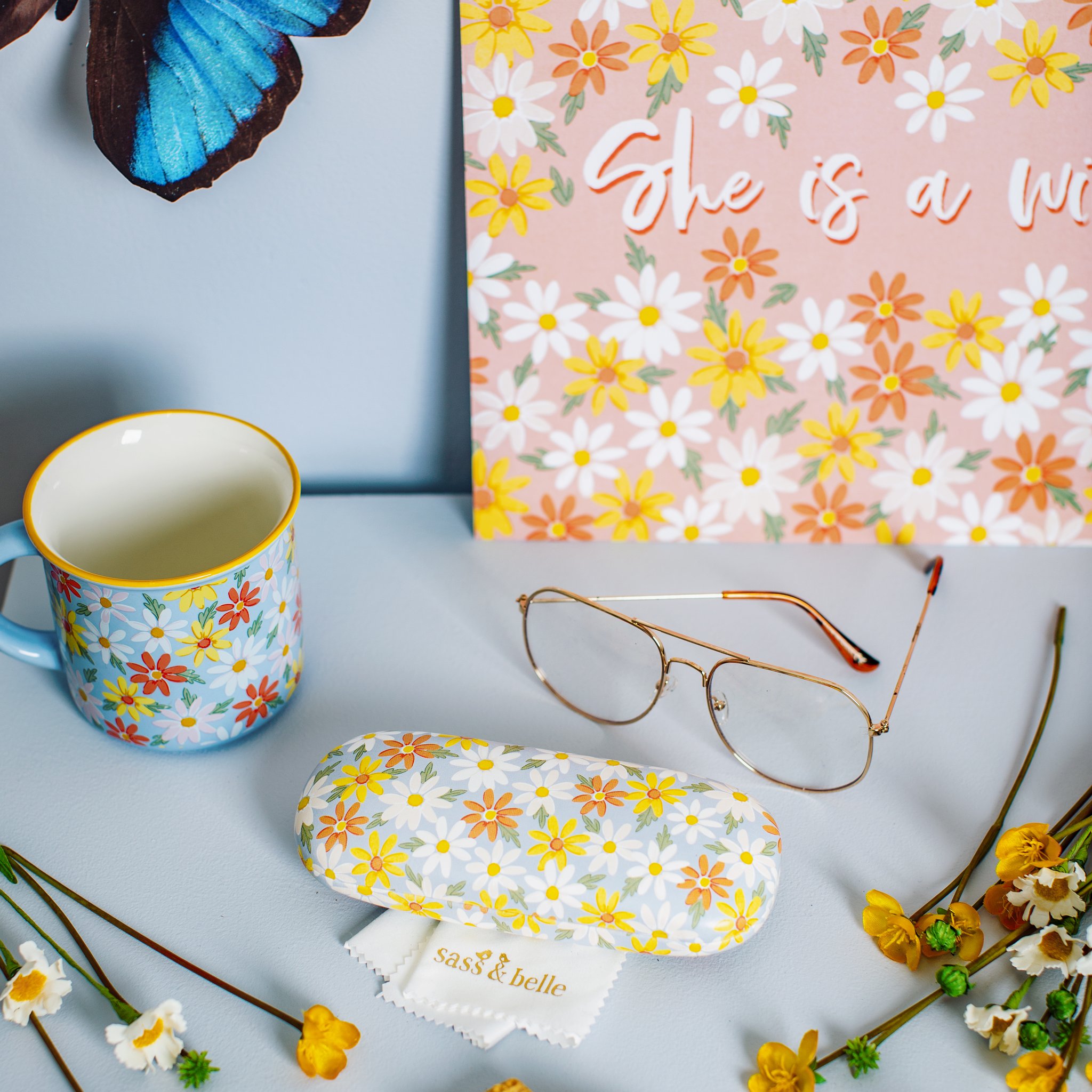 Custodia per occhiali Daisy Sass & Belle con occhiali, panno e tazza floreale su un tavolo azzurro