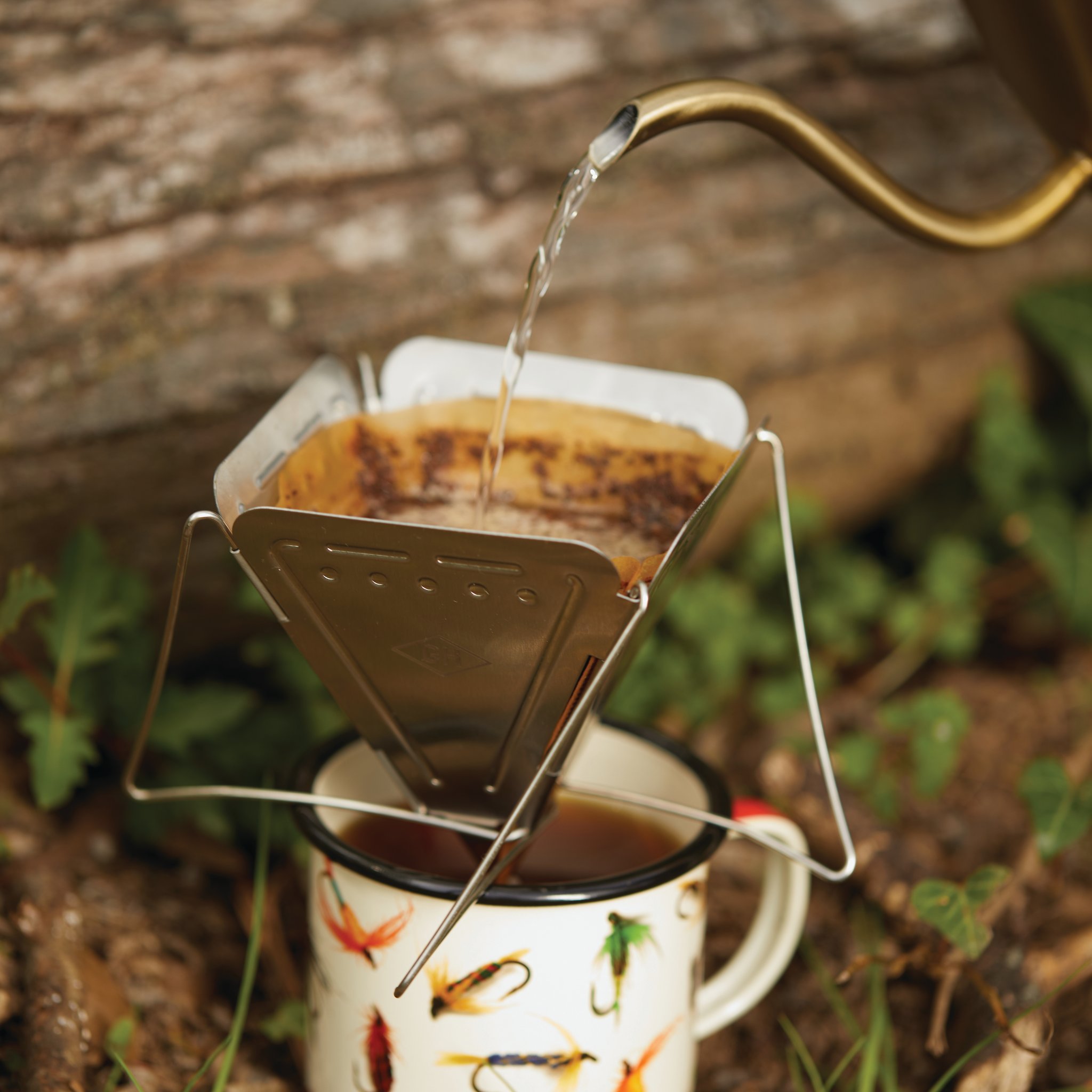 Im Freien giesst jemand Wasser aus einem Kessel auf den Kaffeefilterhalter über einer Emailletasse mit Fliegenmuster