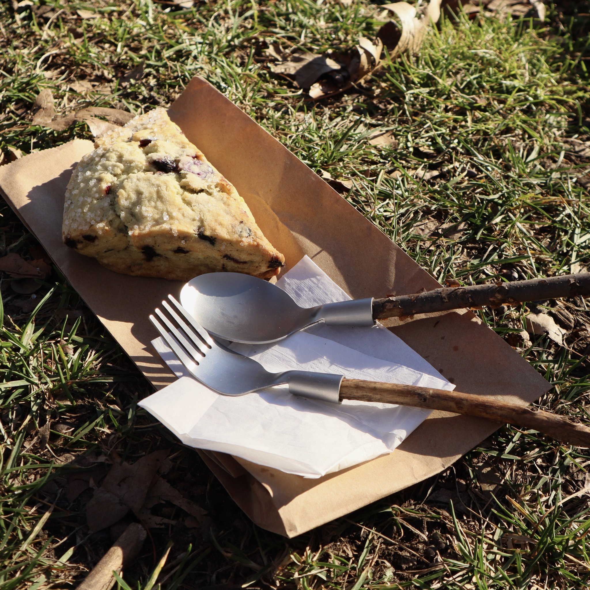 Huckleberry cutlery on paper in the grass next to a scone