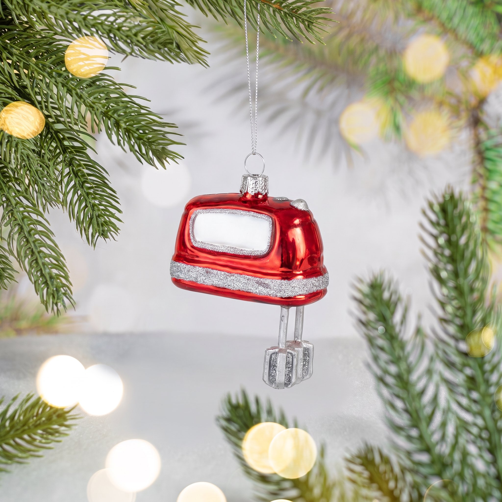 Batteur rouge en verre suspendu à une branche de sapin devant des lumières chaleureuses