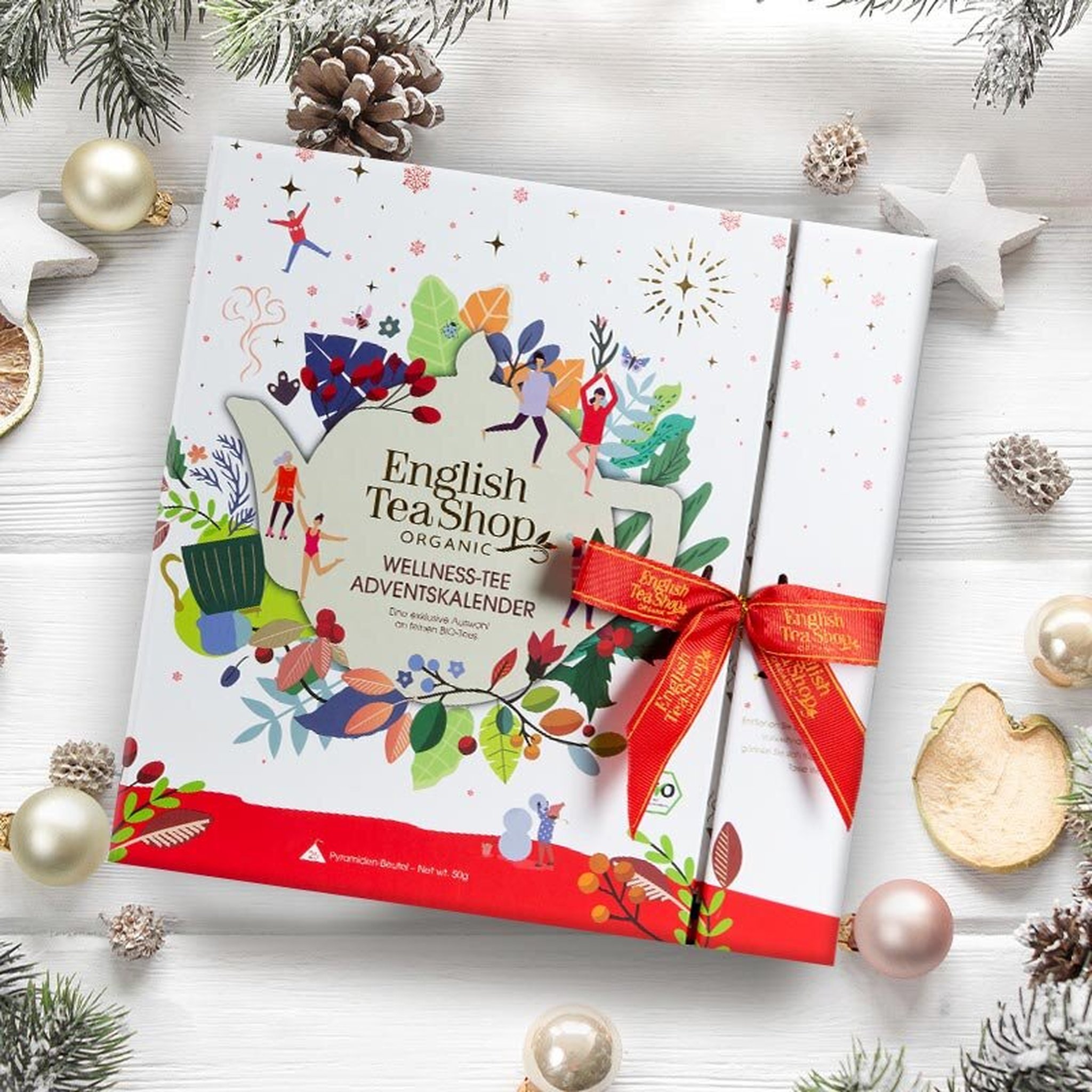 Wellness-Tee-Adventskalender mit roter Schleife zwischen Christbaumkugeln und Tannenzapfen