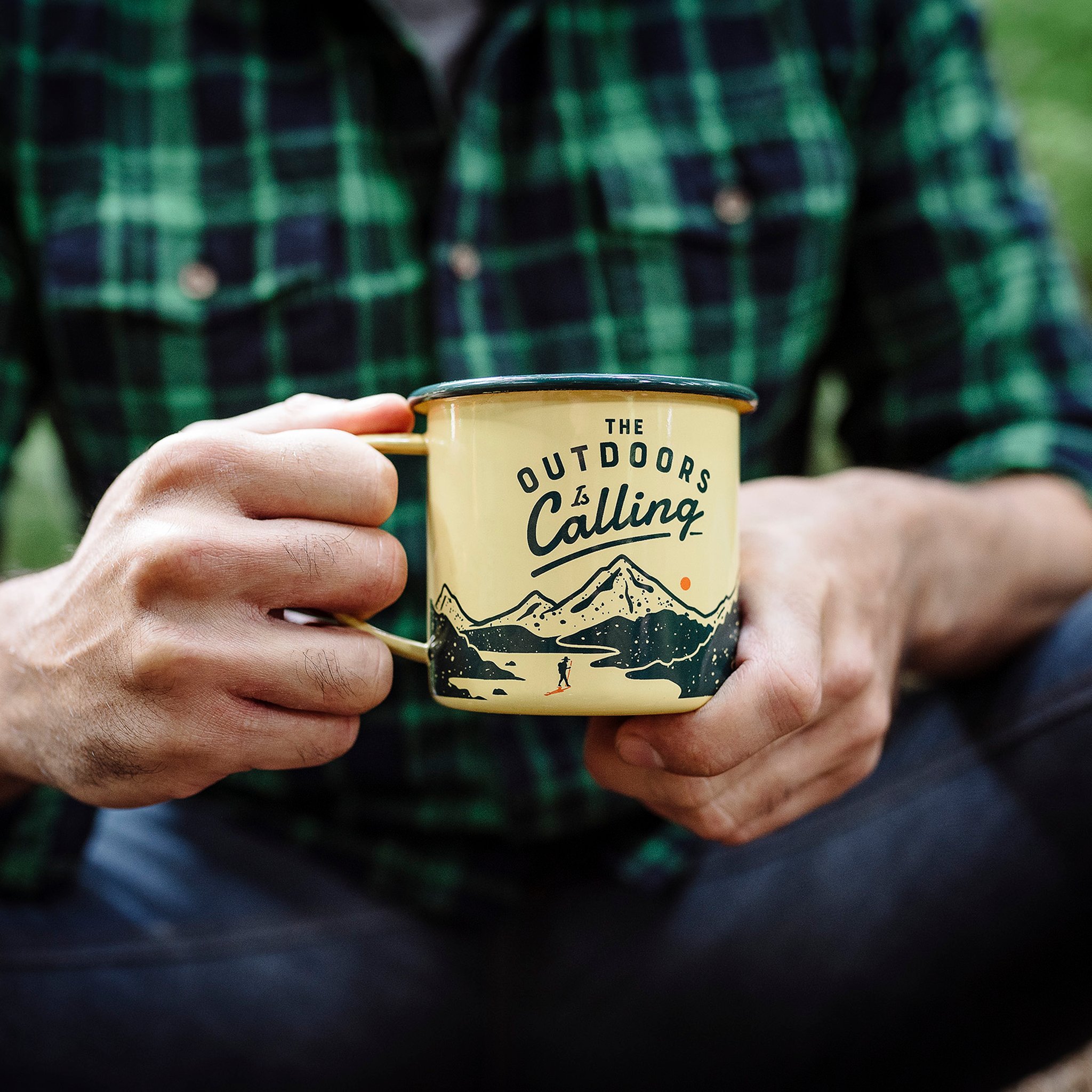 Mann im grün karierten Hemd hält die Outdoor Emaille-Tasse mit beiden Händen vor sich im Freien