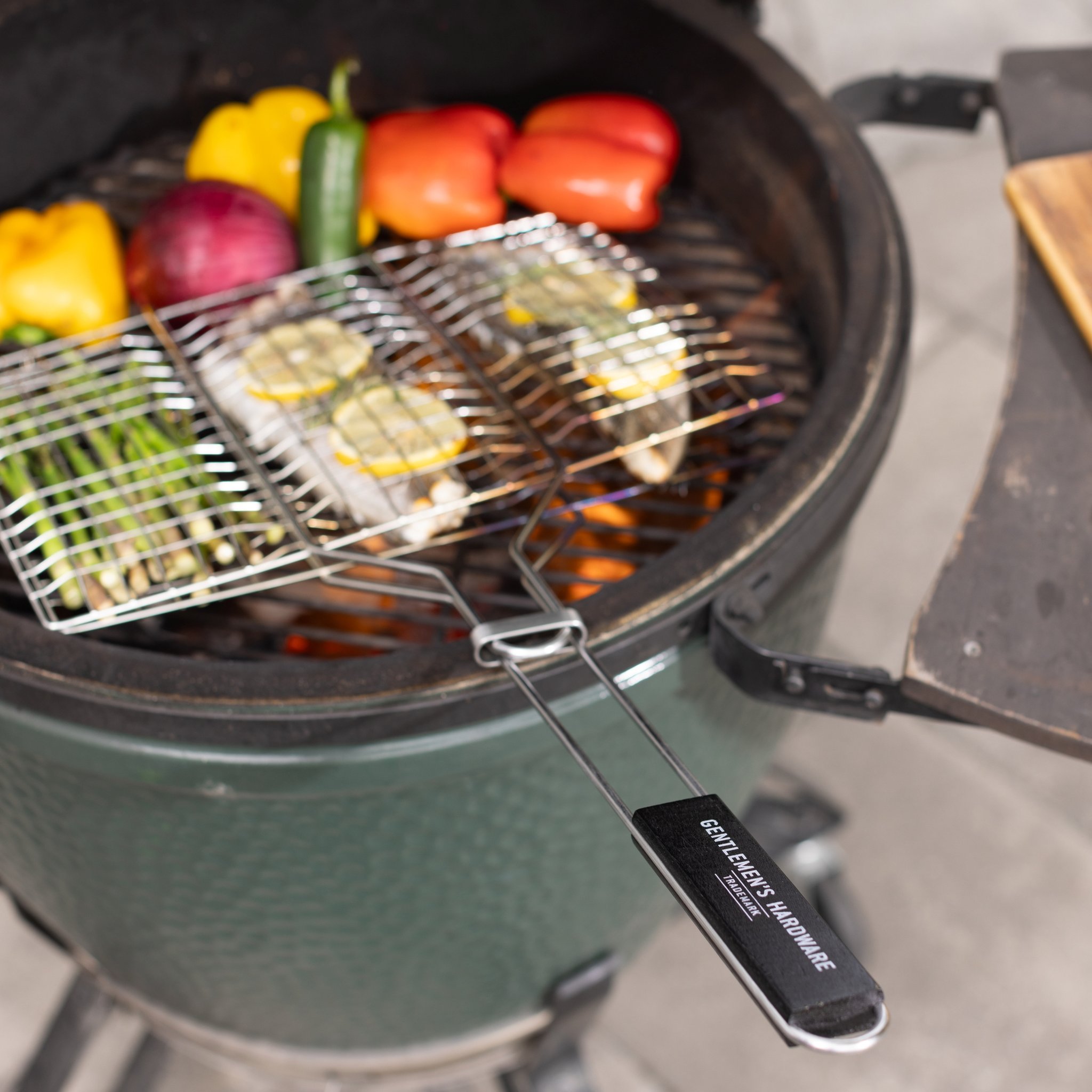 Manico nero del cestino per barbecue in primo piano, con pesce e asparagi sulla griglia verde accanto a un tagliere