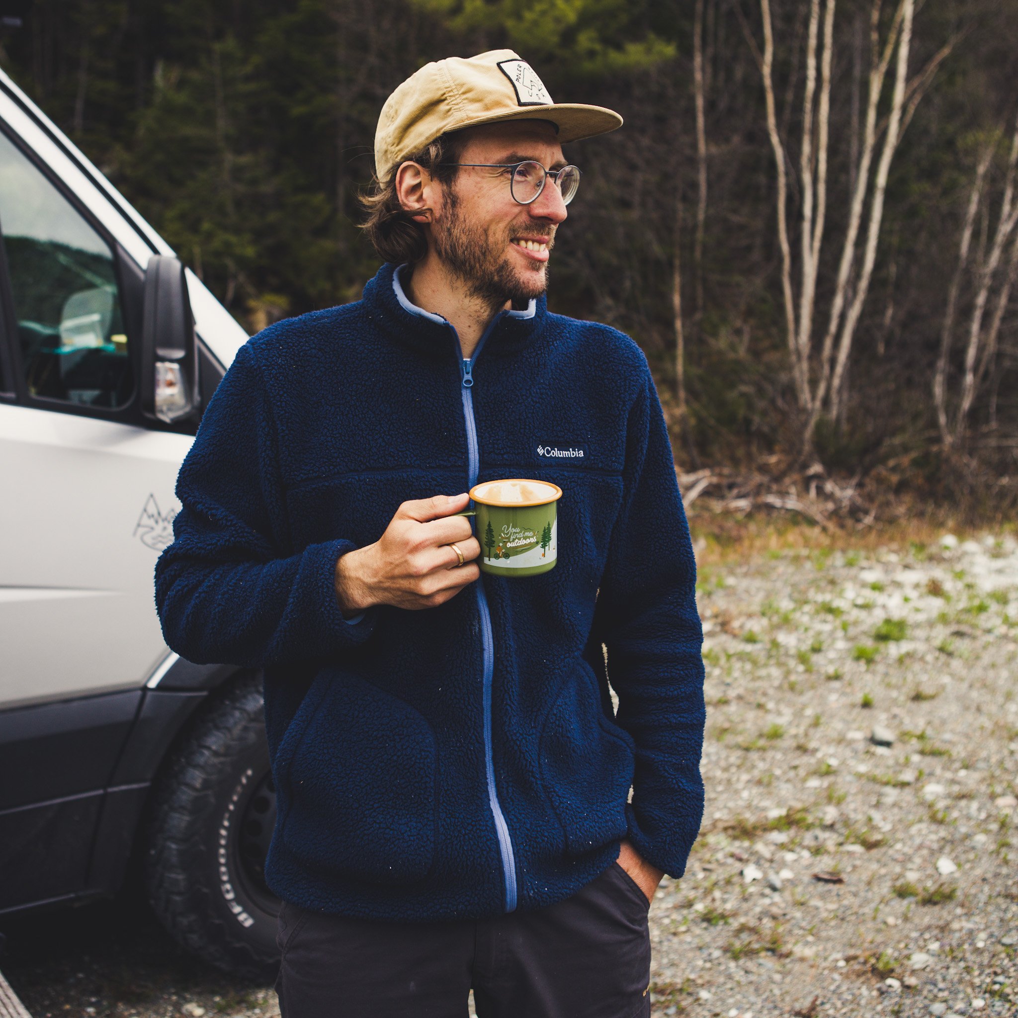 Homme buvant dans la tasse en email olive You find me Outdoors devant un van