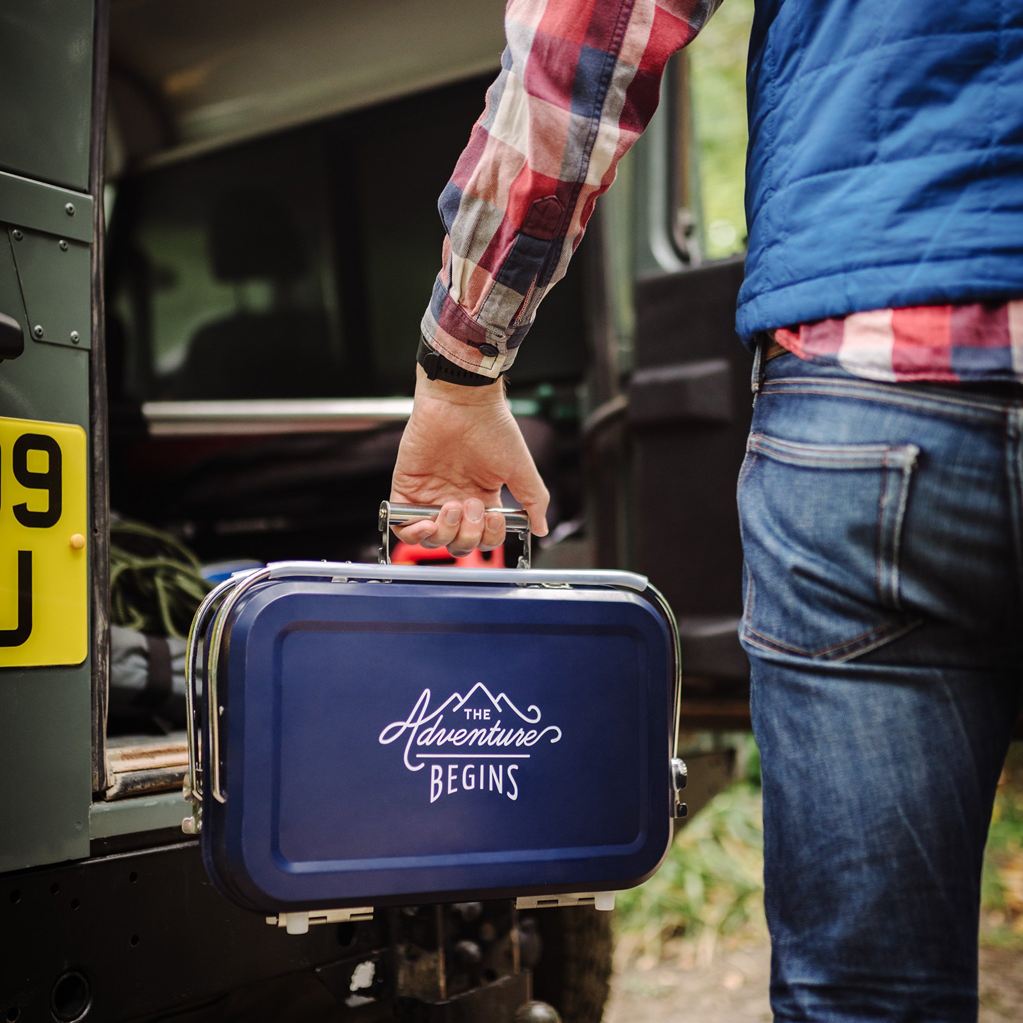 Person in jeans carrying the closed Large Portable BBQ by its handle like a suitcase, with a green off-road vehicle behind