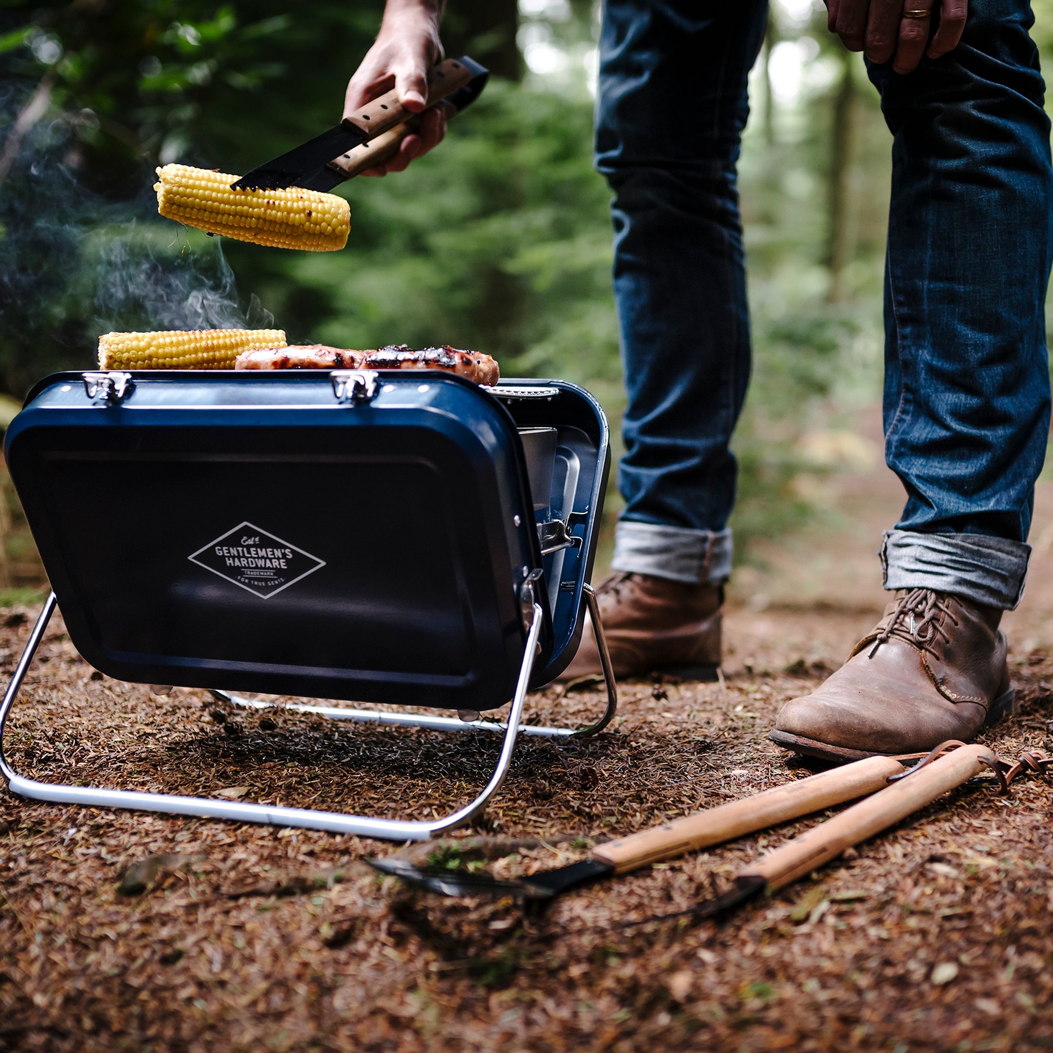 Grilling in the woods: tongs lift a corn cob while more corn and sausages cook on the Large Portable BBQ