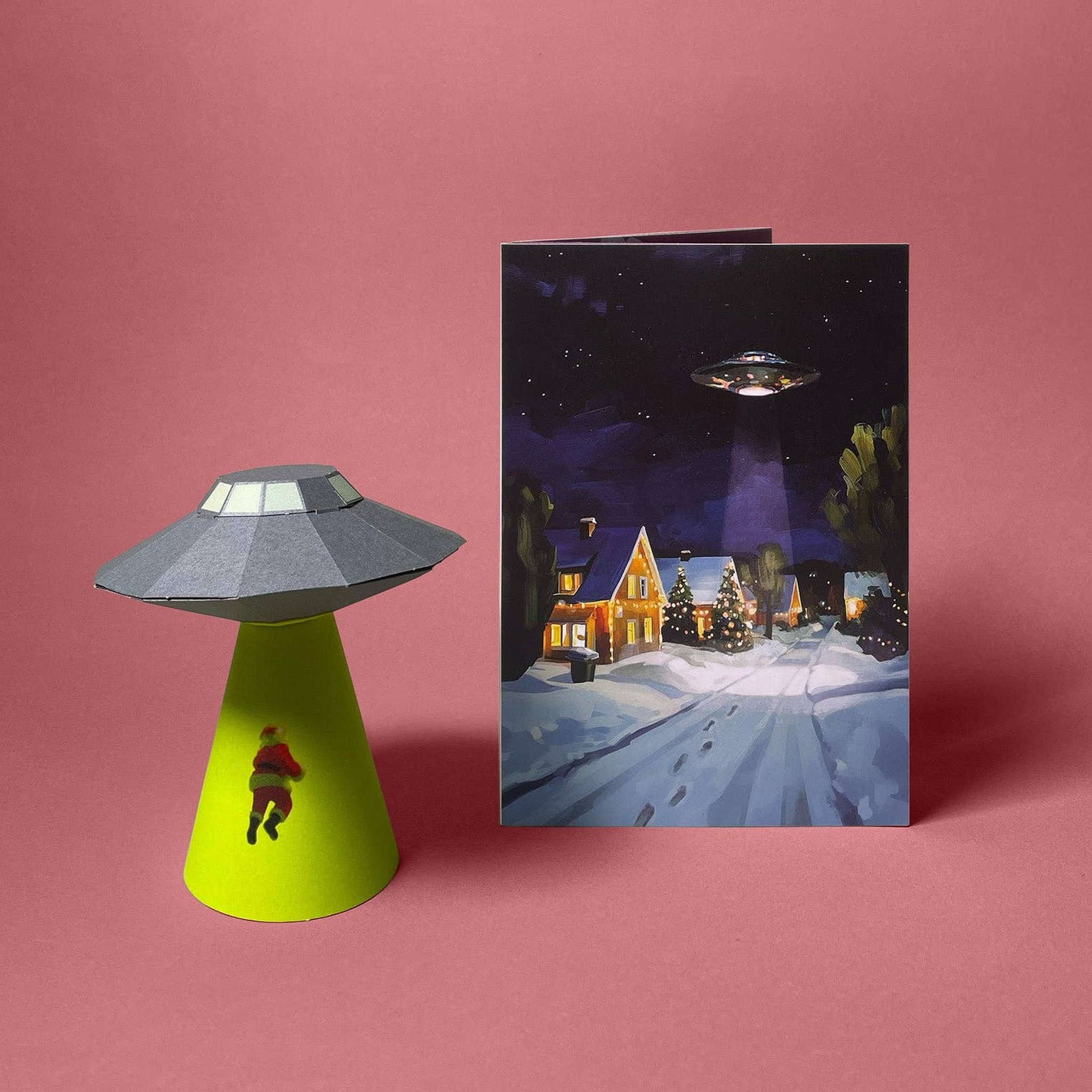 Gefaltetes 3D-Modell UFO, das den Weihnachtsmann entführt neben der Weihnachtskarte mit UFO über verschneitem Dorf von Faltmeister