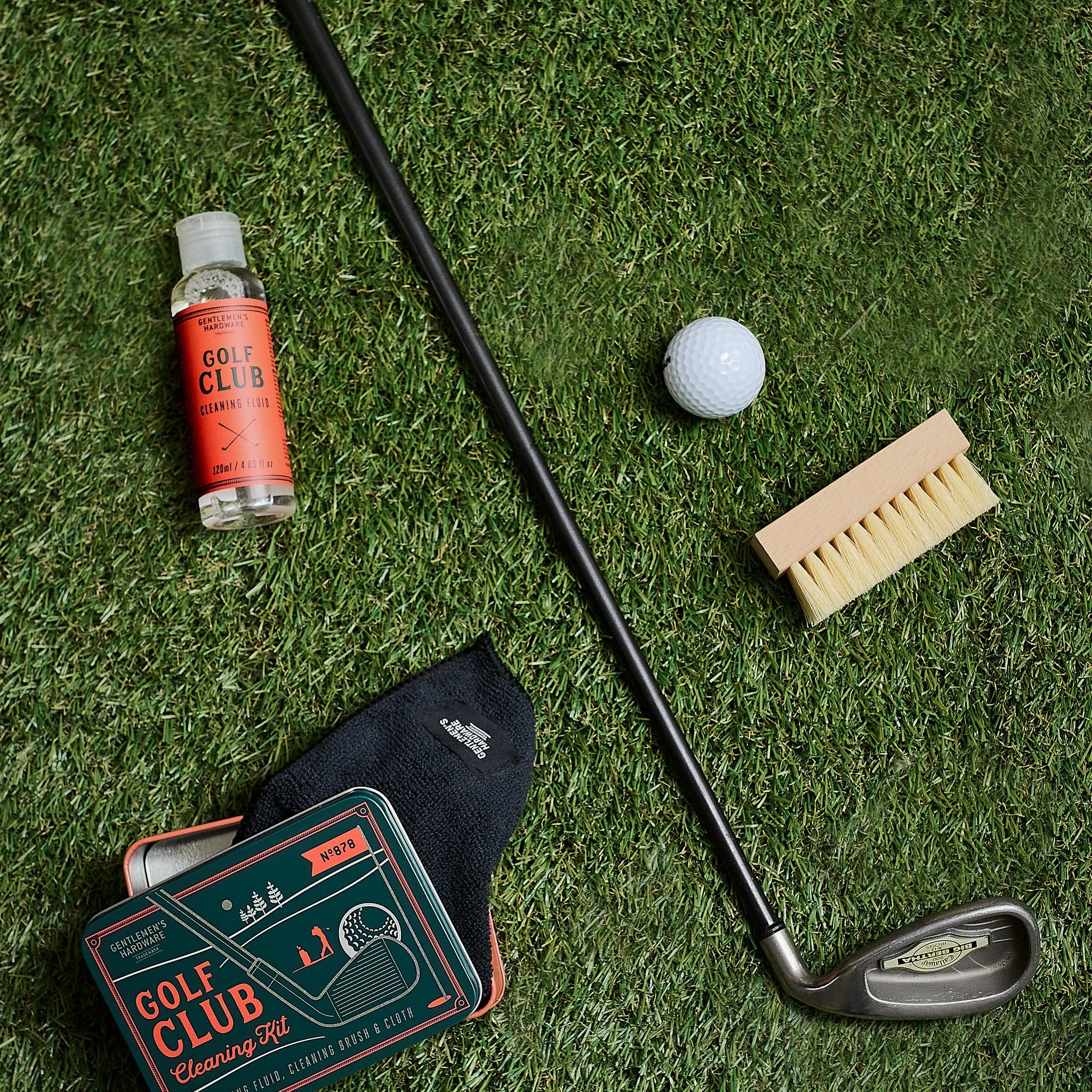 On the grass lie a golf club, ball, brush and bottle, with the black cloth peeking out from under the cleaning set tin