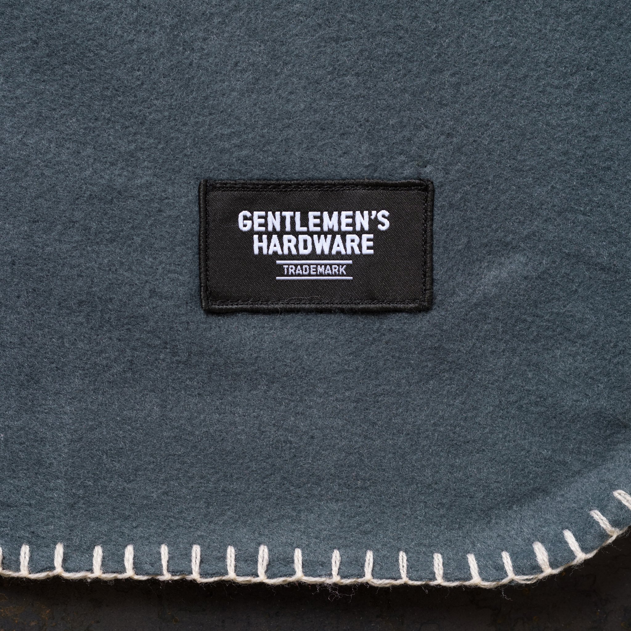 Nahaufnahme der Campingdecke in Grau mit aufgenähtem schwarzem Label und heller Ziernaht am unteren Rand