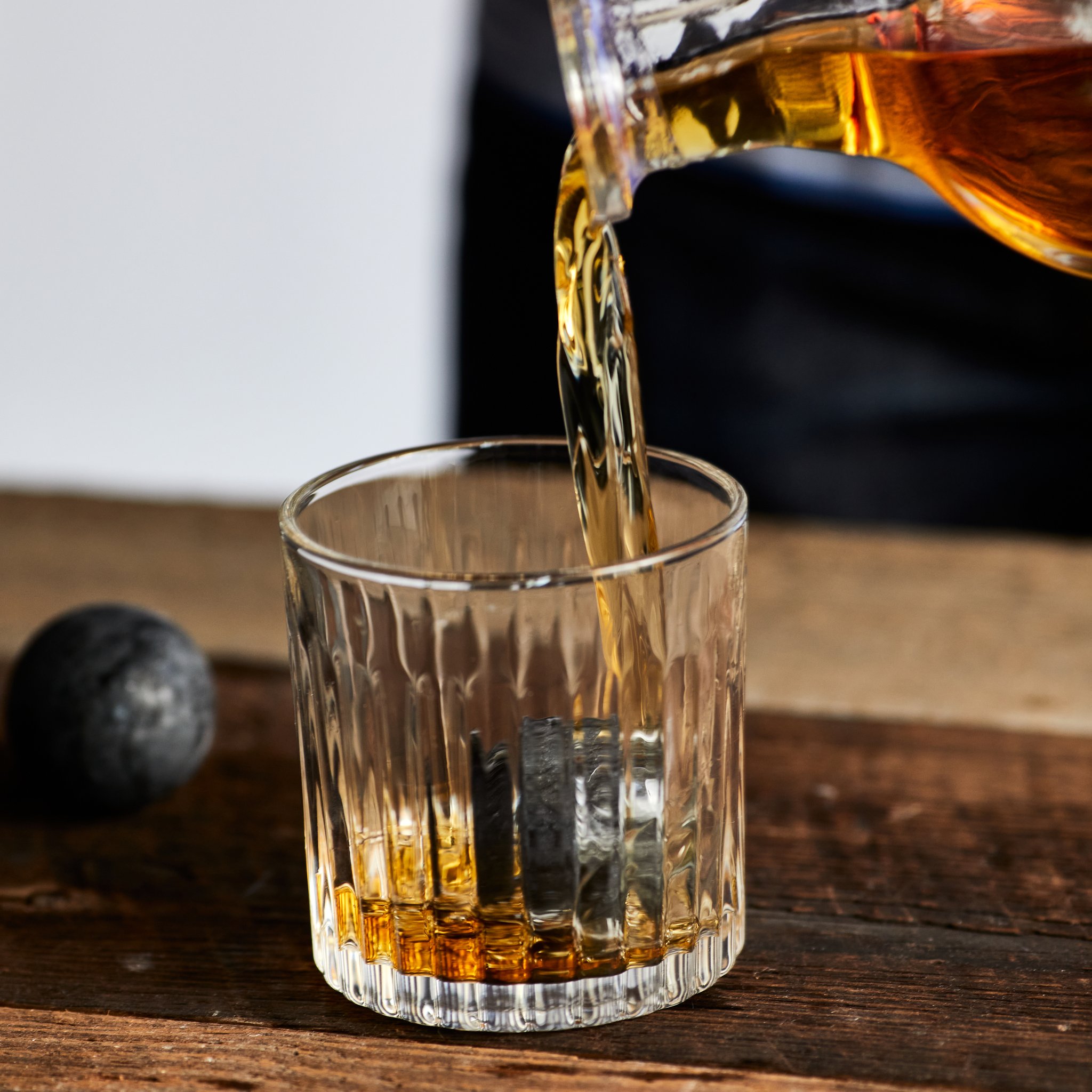 Whisky wird aus einer Karaffe in ein geriffeltes Glas auf einem Holztisch gegossen, daneben liegt eine Granitkugel