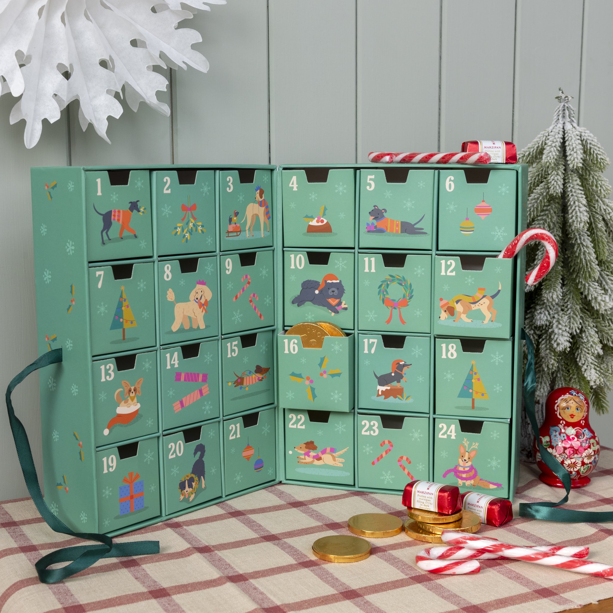 Aufgeklappter Festive Friends Adventskalender vor Weihnachtsdeko mit Schneeflocke und Tannenbaum