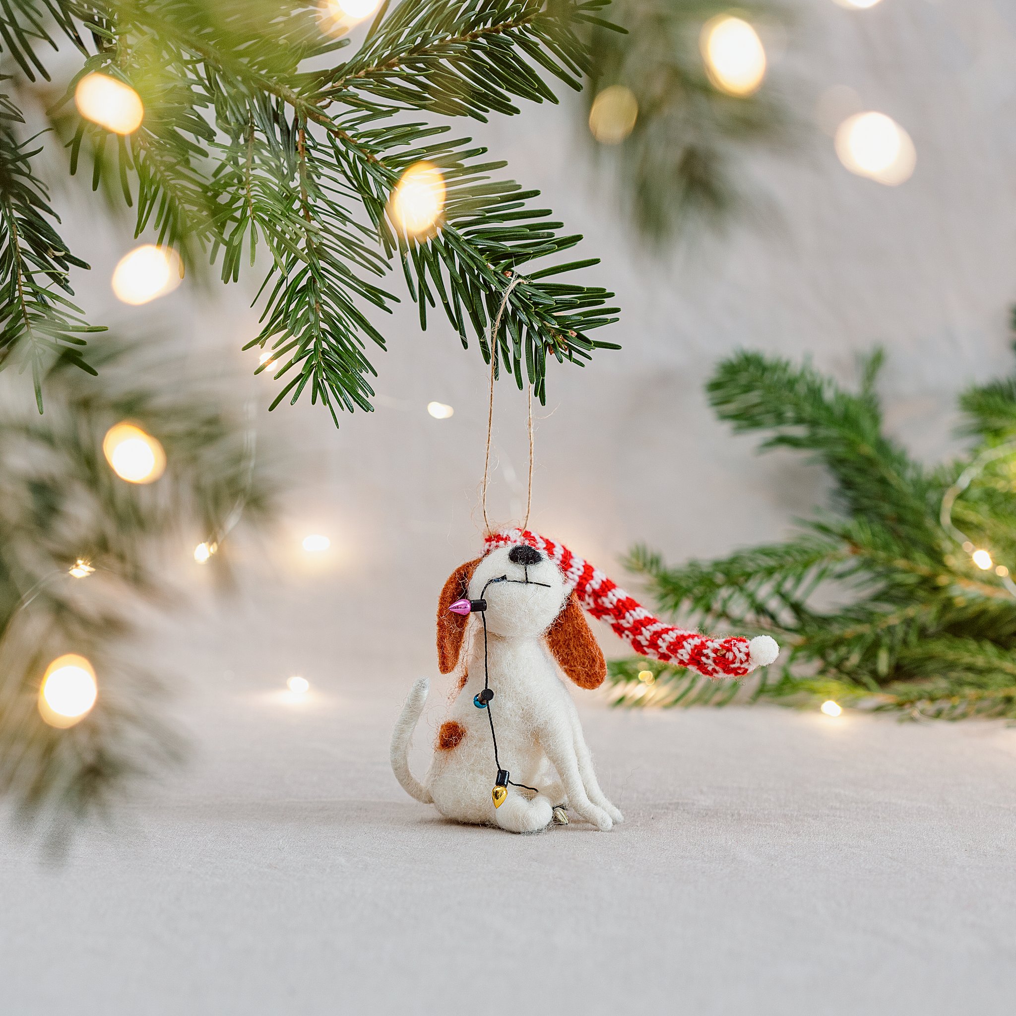 Felt dog ornament hanging from a fir branch with warm lights behind