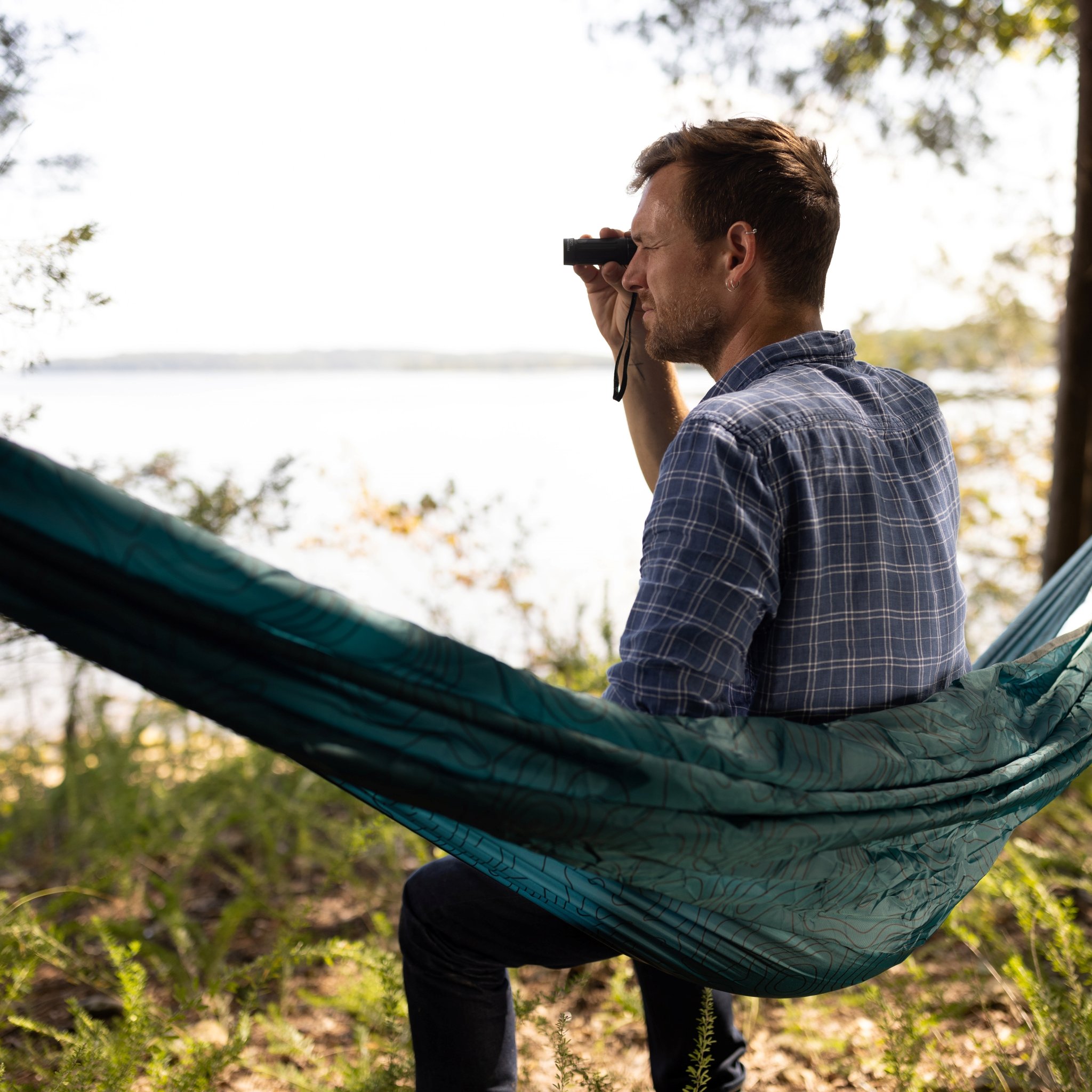Homme assis dans un hamac au bord du lac, regardant au loin avec le monoculaire du Kit d'exploration