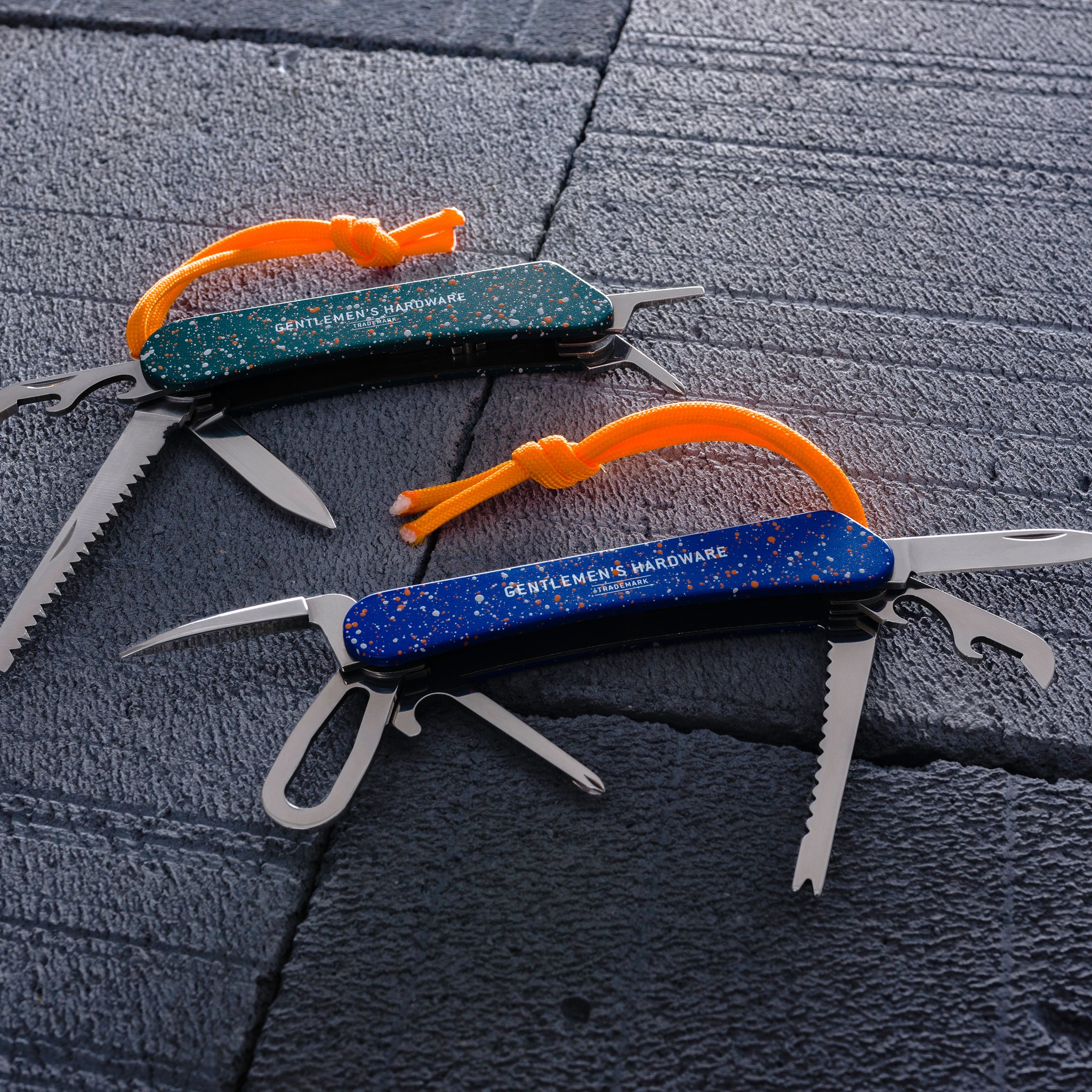 Zwei aufgeklappte Wildnis Multitools in Grün und Blau mit orangen Bändern auf grauem Steinboden