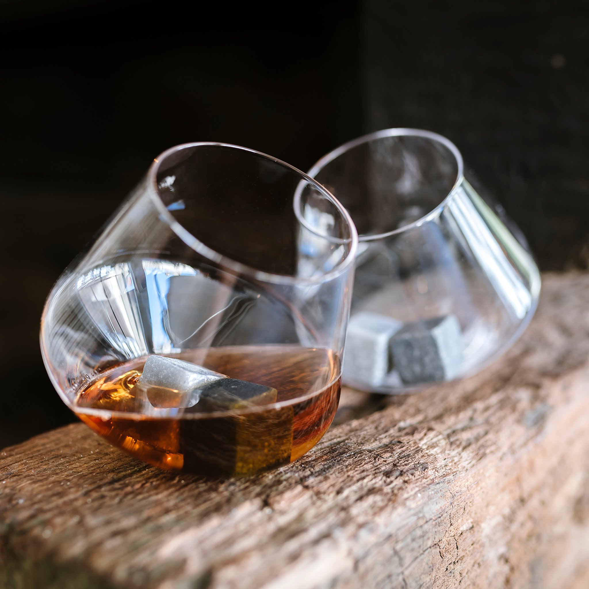Two tilted Rocking Whisky Glasses with whisky and chilling stones resting on a rustic wooden beam