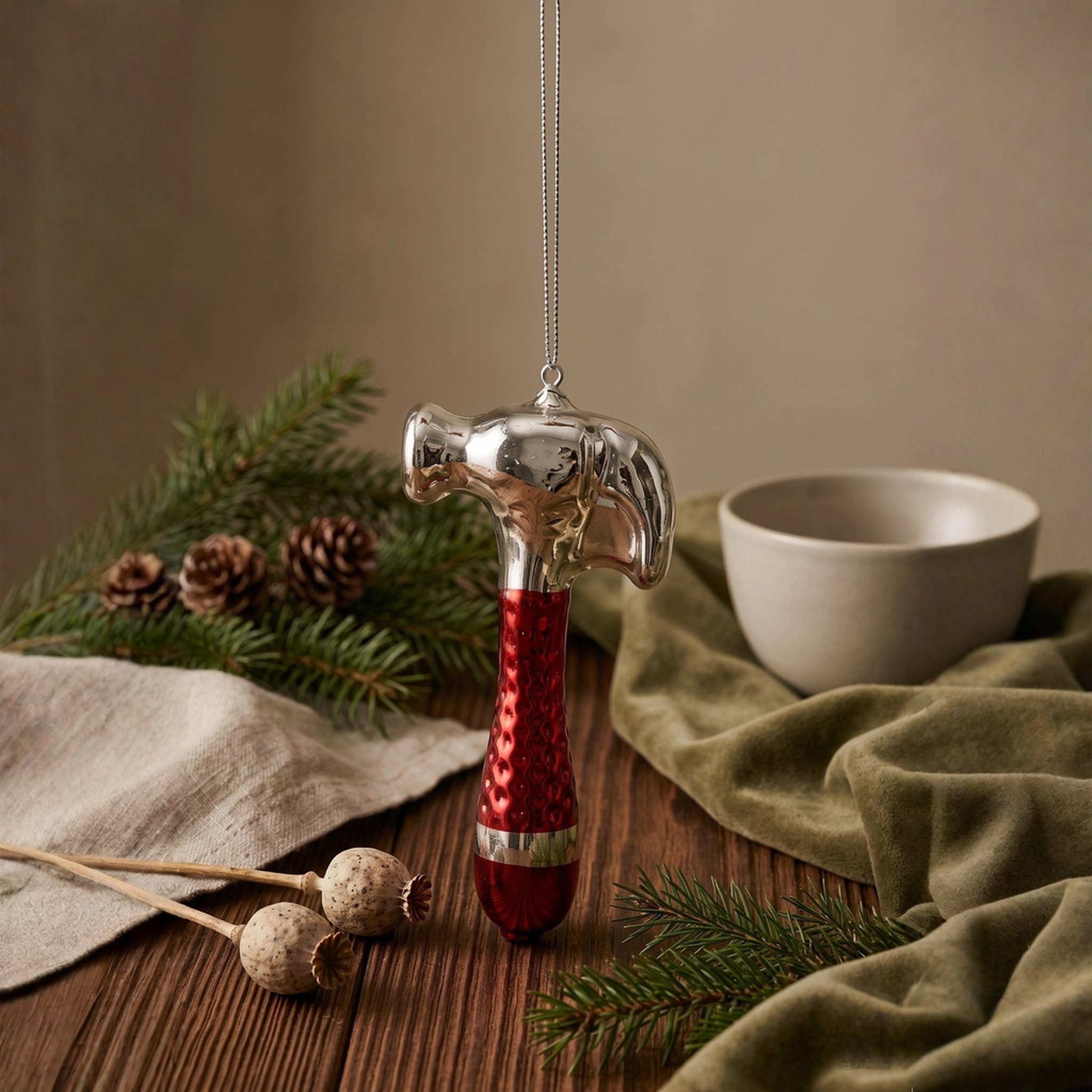 Christbaumschmuck Hammer aus Glas über einem Holztisch mit Tannenzweigen, Mohnkapseln und Schale