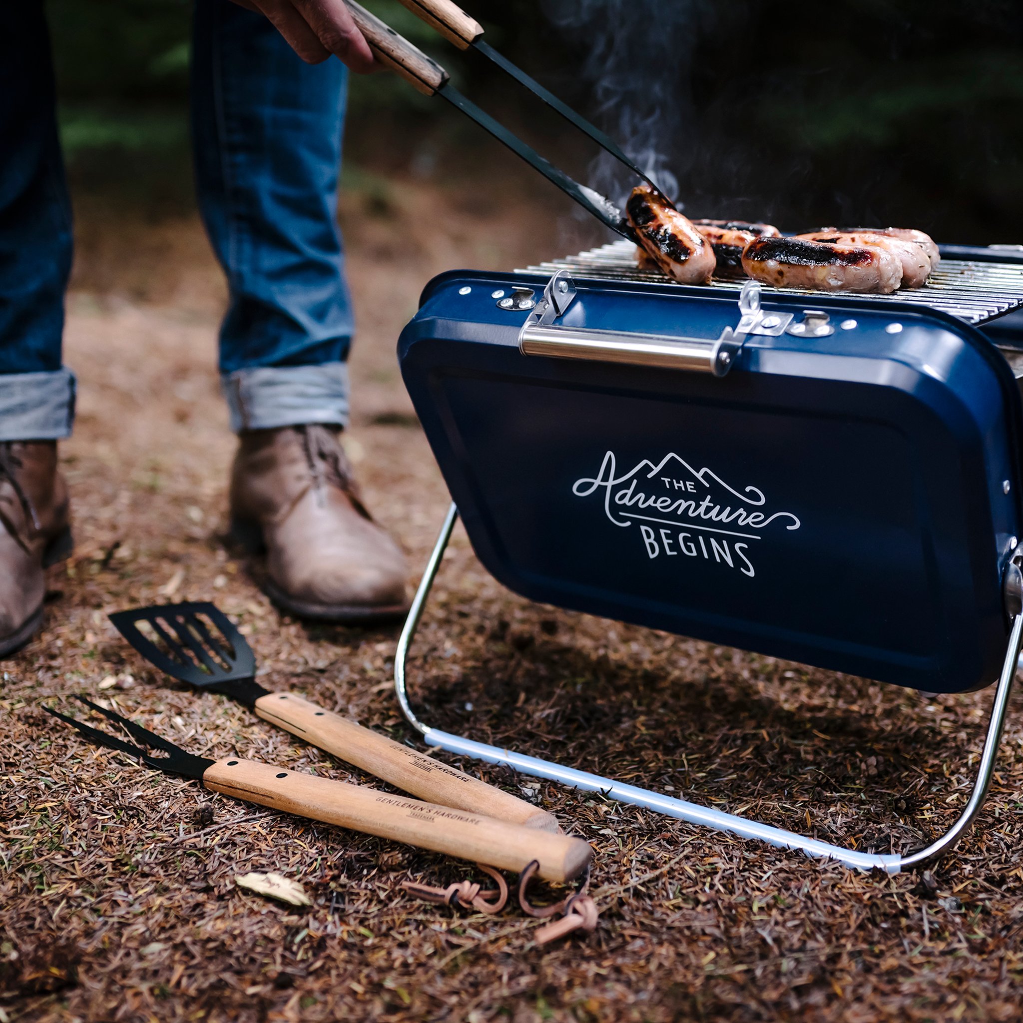 Grilled sausages on the rack of the Large Portable BBQ, with a spatula and fork with wooden handles on the forest floor