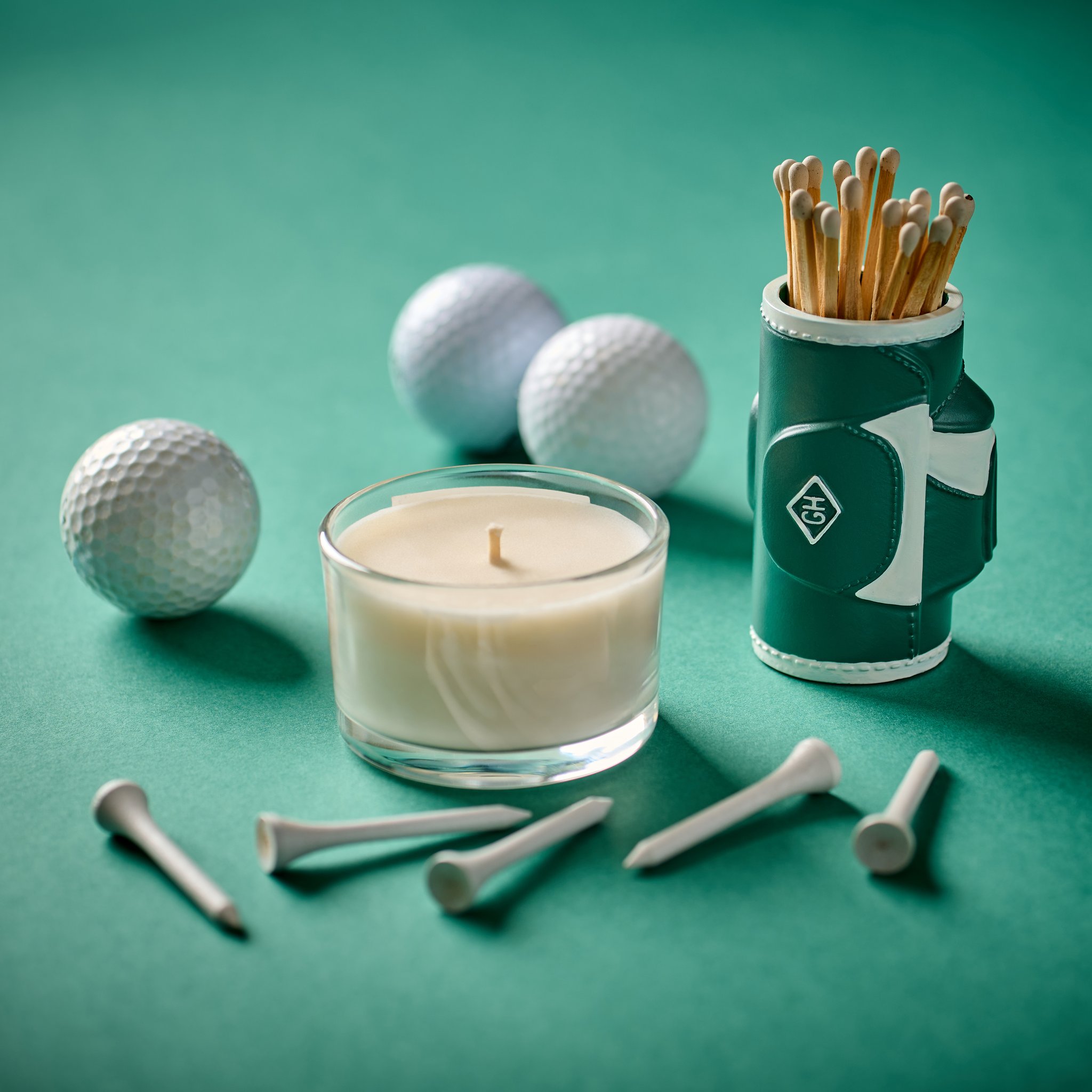 Portafiammiferi sacca da golf accanto a una candela in vetro, palline e tee da golf su fondo verde