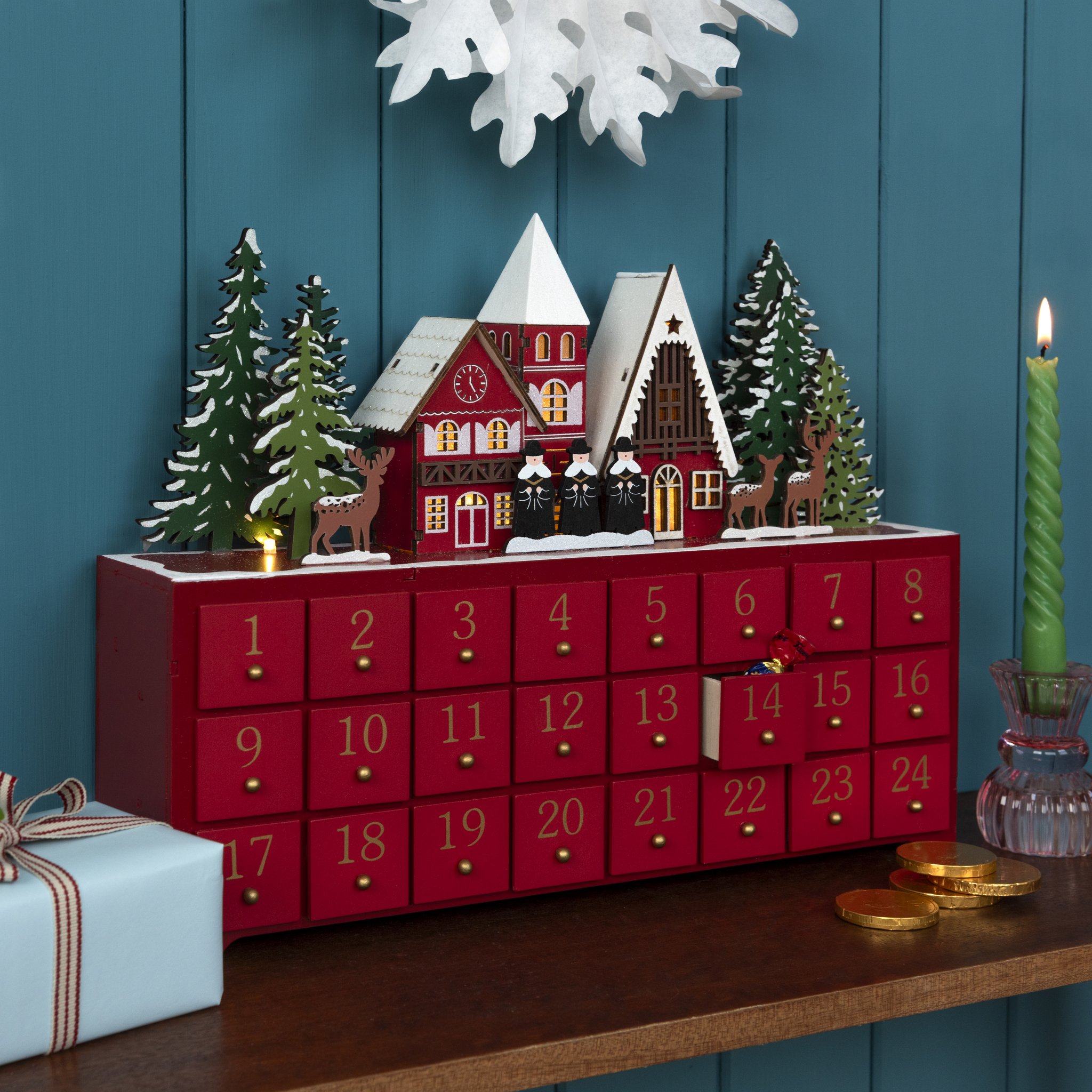 Weihnachtssinger Adventskalender aus Holz mit beleuchtetem Dorf auf der Kommode neben Geschenk