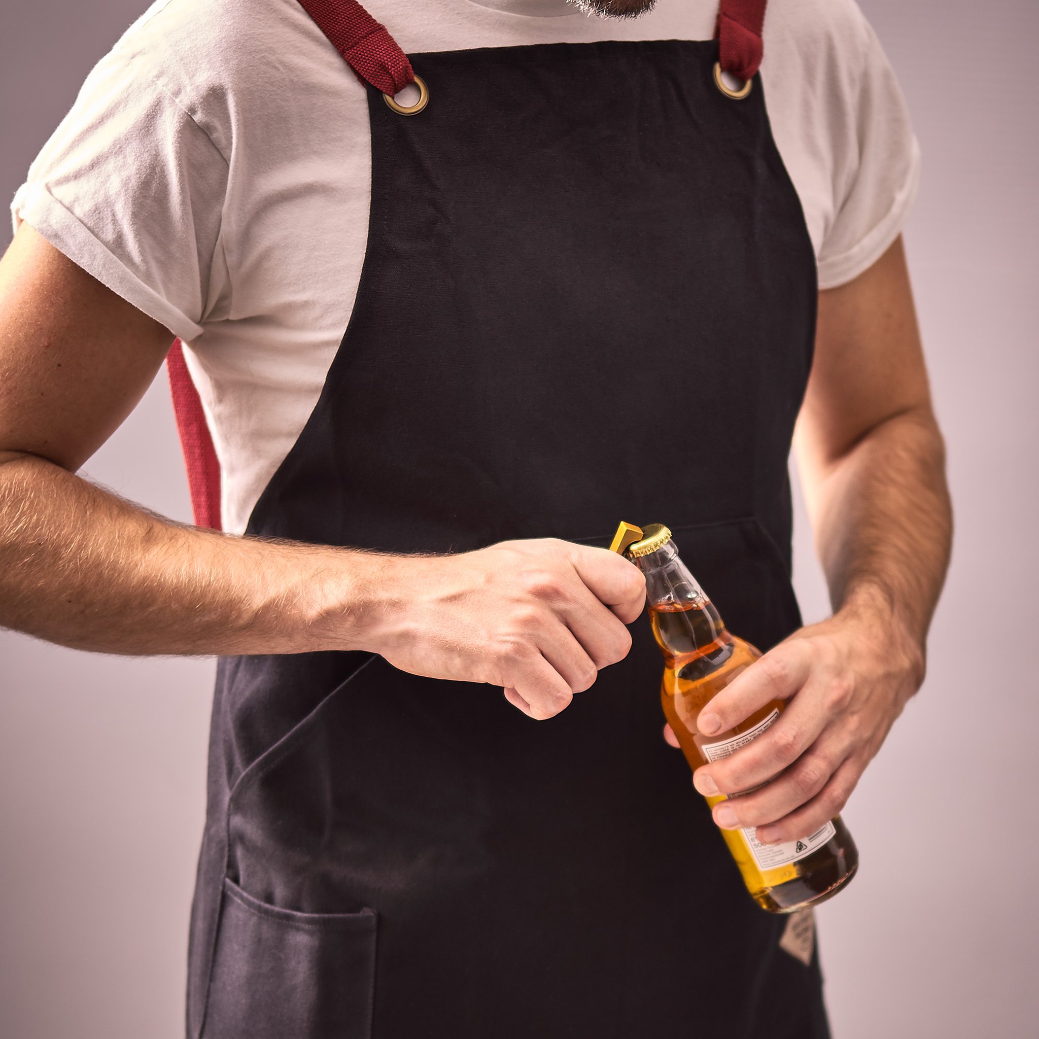 Man wearing the black BBQ apron opens a bottle of beer with the bottle opener held in his hand