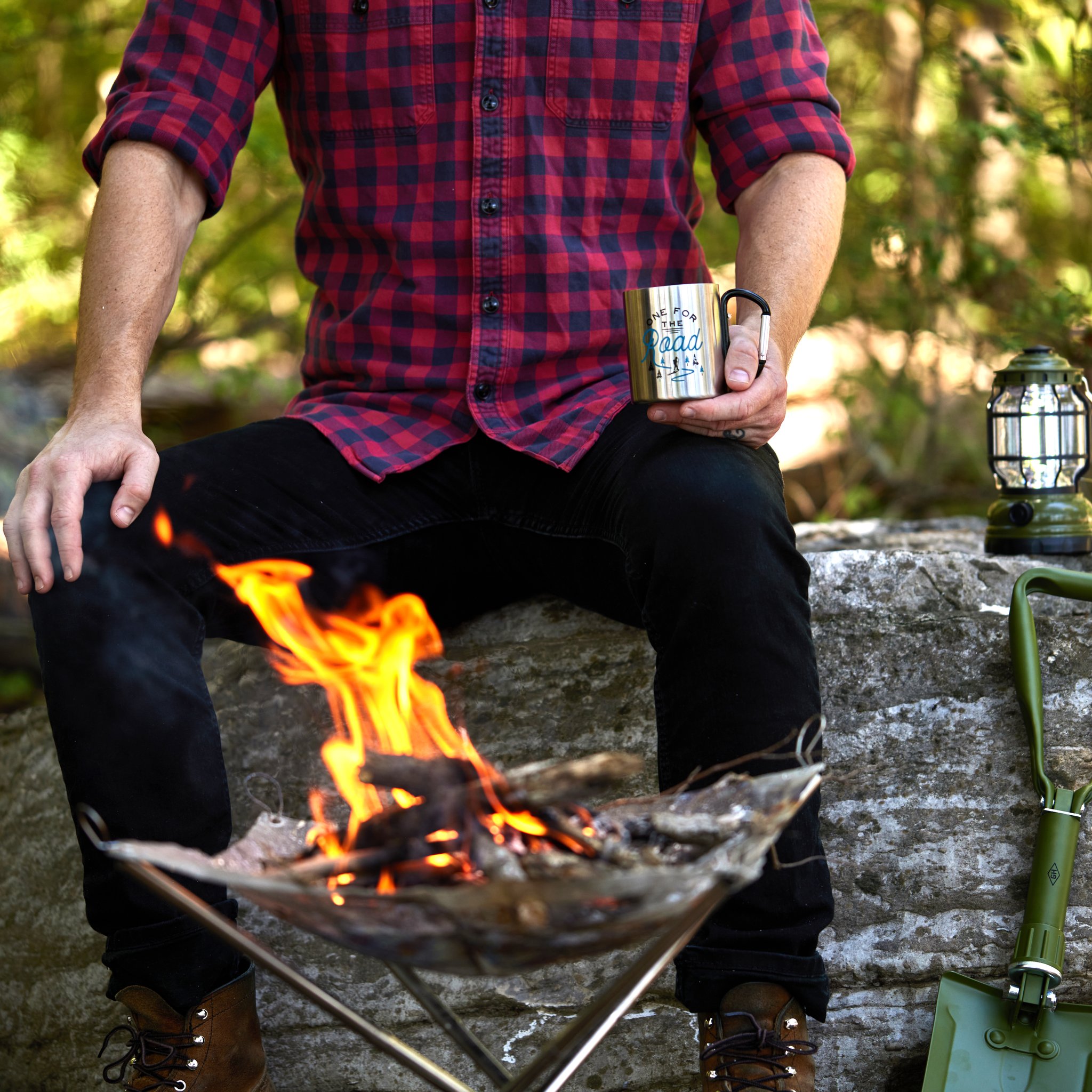 Man in a checked shirt sits by a campfire holding a metal mug, the folding shovel propped against a rock beside him