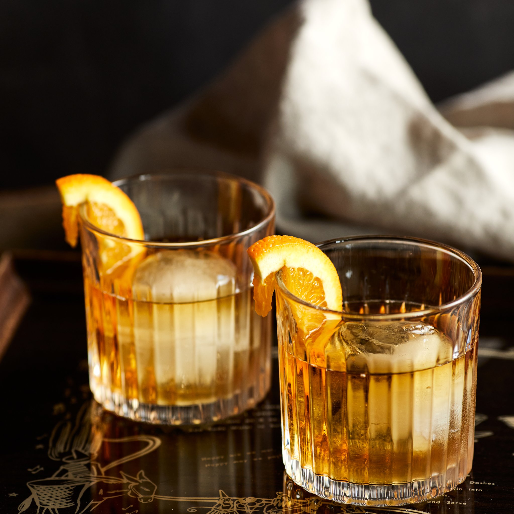 Zwei Gläser mit Whisky, Eiskugel und Orangenscheibe auf dunklem Untergrund, im Hintergrund ein helles Tuch