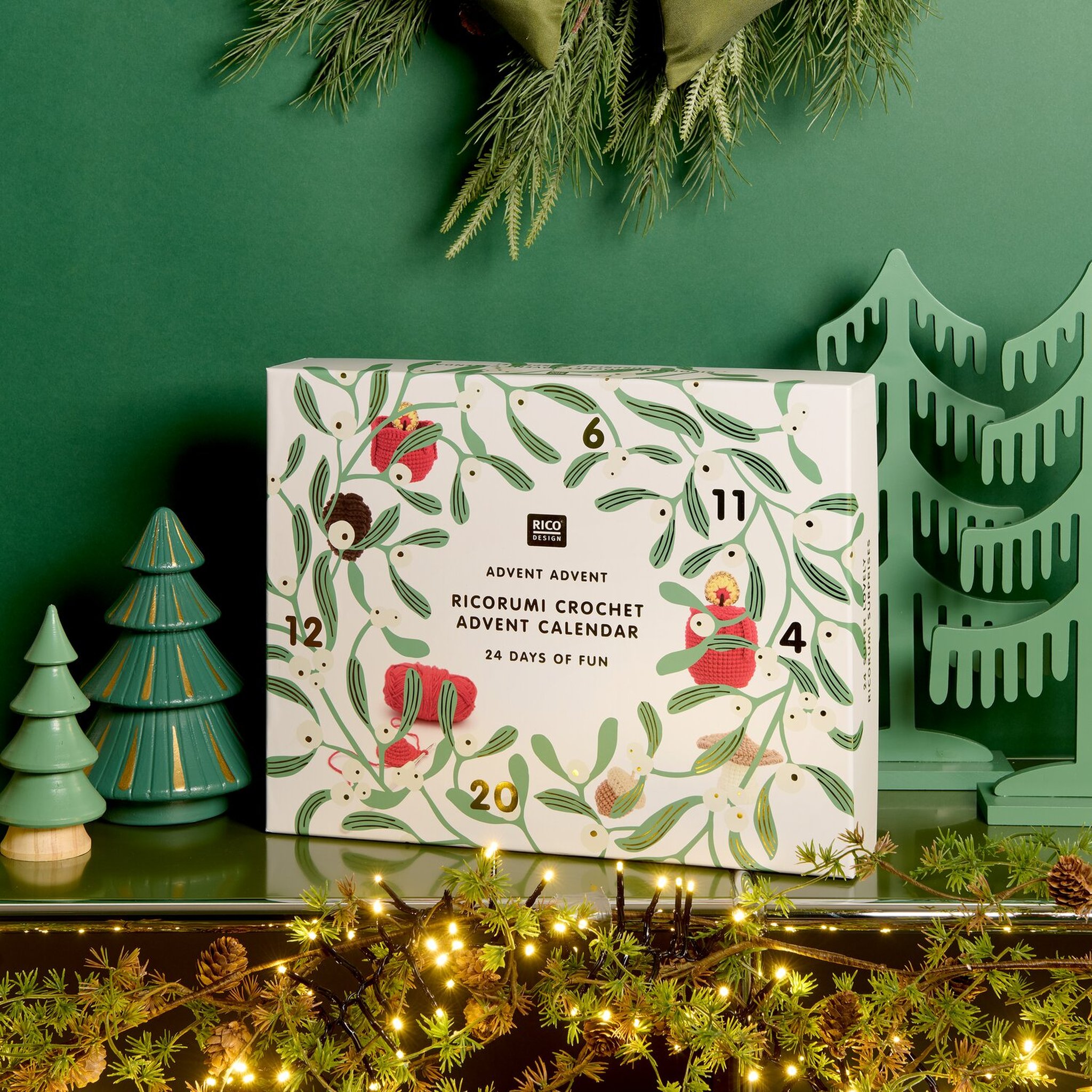 Ricorumi Adventskalender auf einer Kommode vor grüner Wand, umgeben von Tannenbäumchen und Lichterkette
