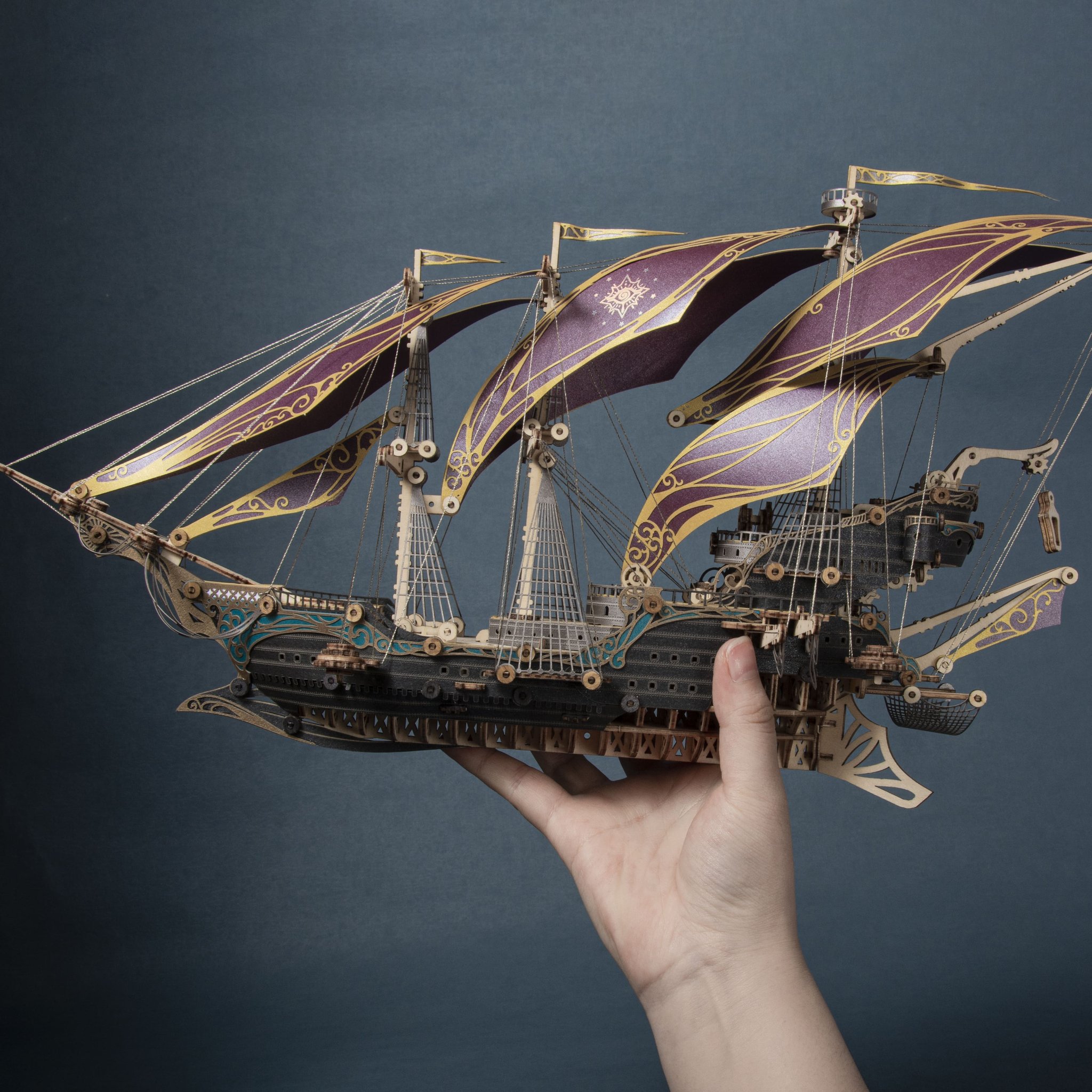 A hand holds the built wooden model with purple paper sails against a dark background