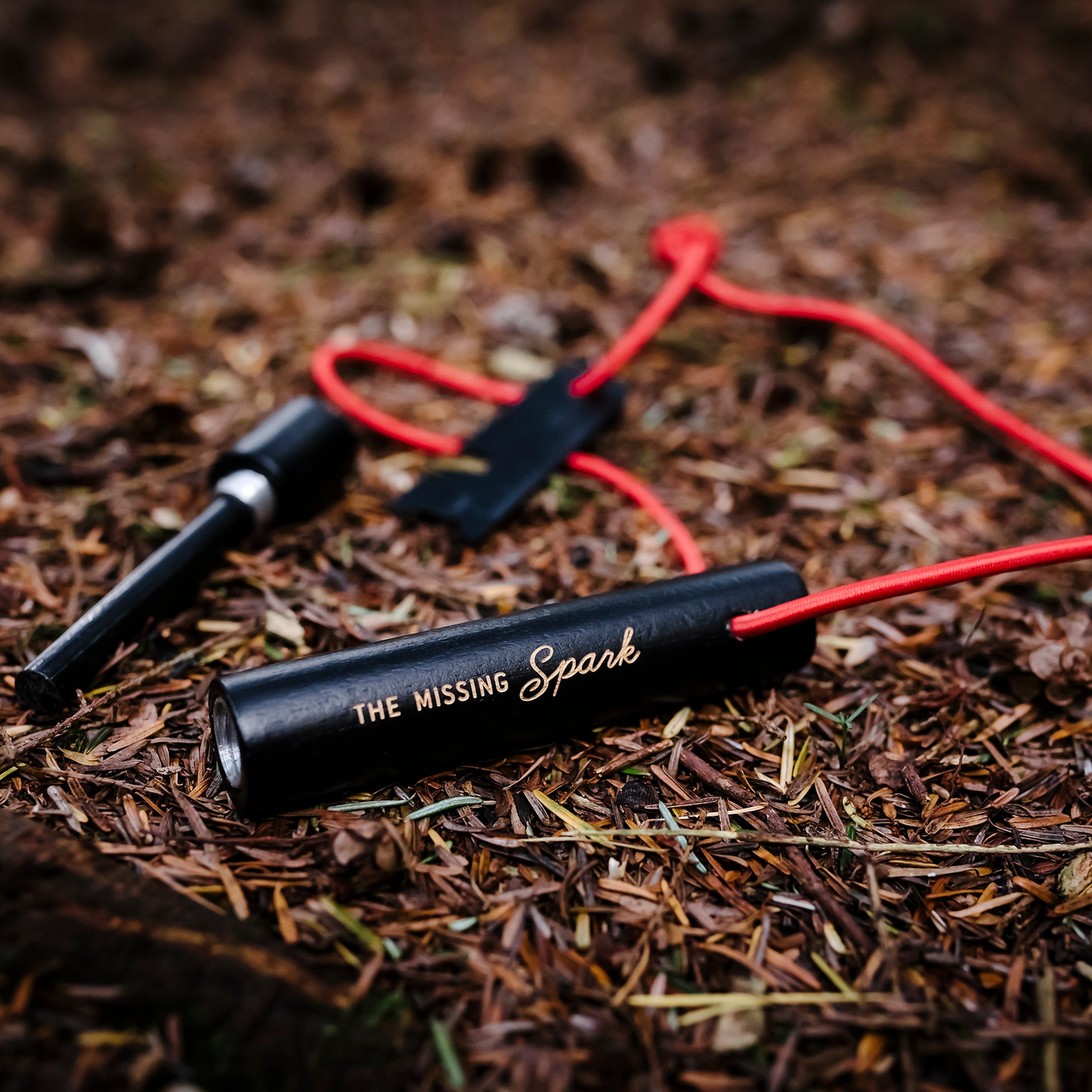 Fire Starter with red cord and striker lying on a forest floor, engraved with the words «The Missing Spark»