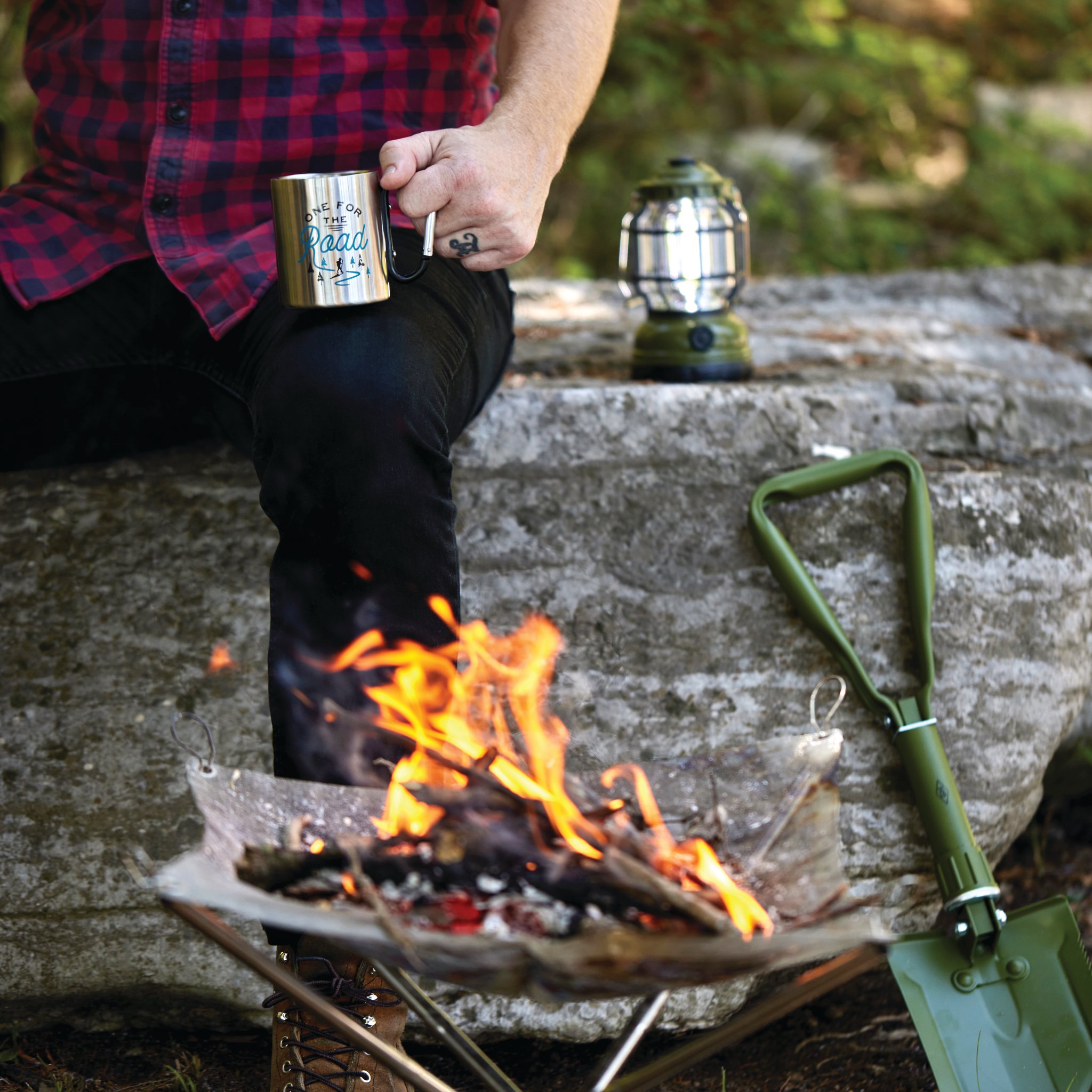 Flames in a fire pit before a large rock, with a lantern, a mug held in one hand and the folding shovel leaning beside it