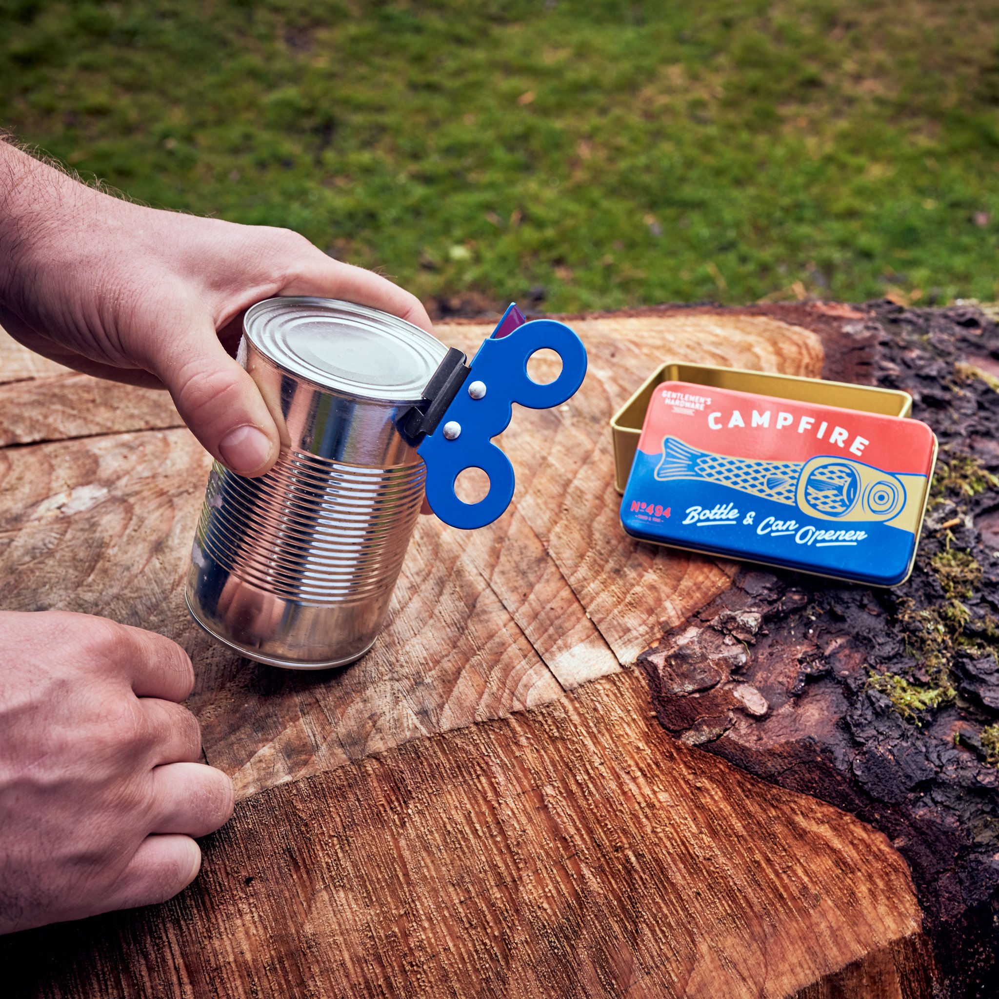 Hand opening a tin can with the Campfire opener on a tree stump outdoors, the gift tin resting beside it