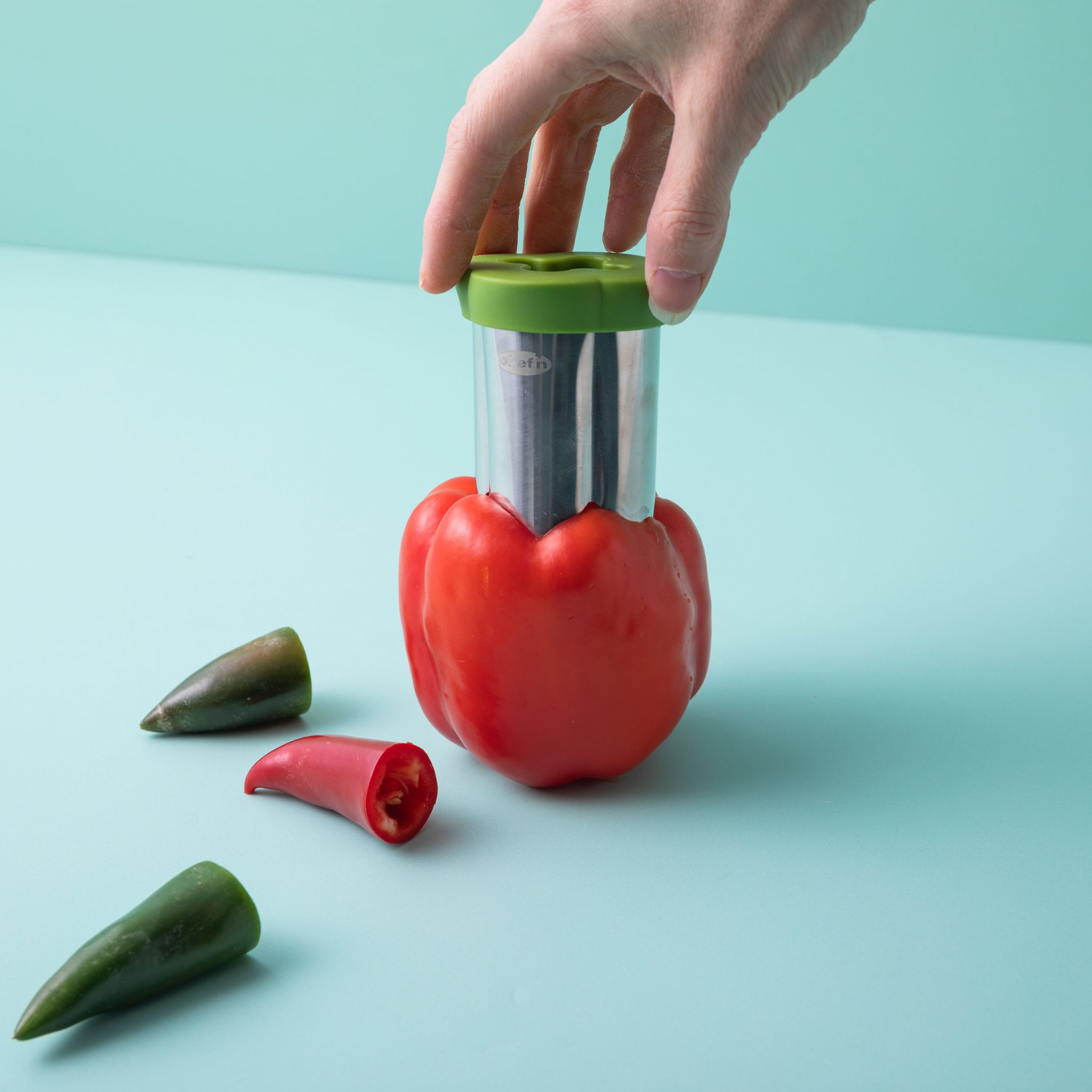 Paprika-Entkerner steckt in roter Paprika, daneben Chilischoten auf türkisem Grund