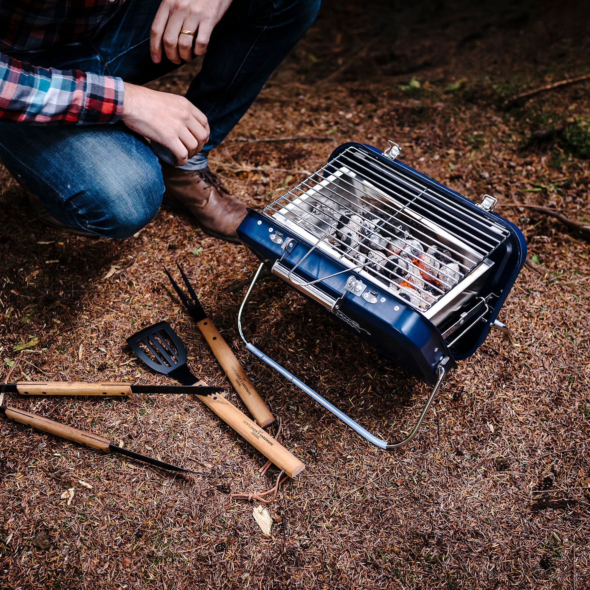 View from above of the Large Portable BBQ with glowing charcoal under the rack, a crouching man and grill tools beside it