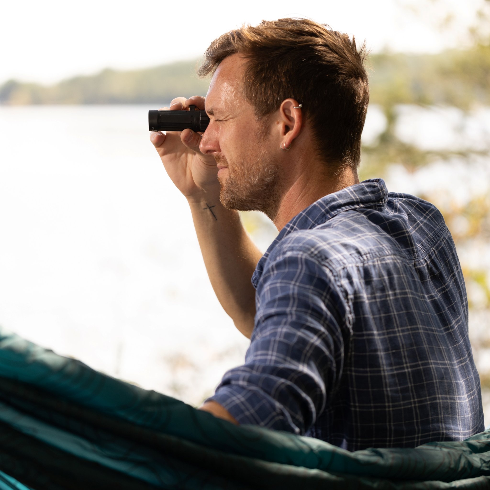 Homme en chemise à carreaux observant un lac avec le monoculaire du Kit d'exploration, hamac au premier plan
