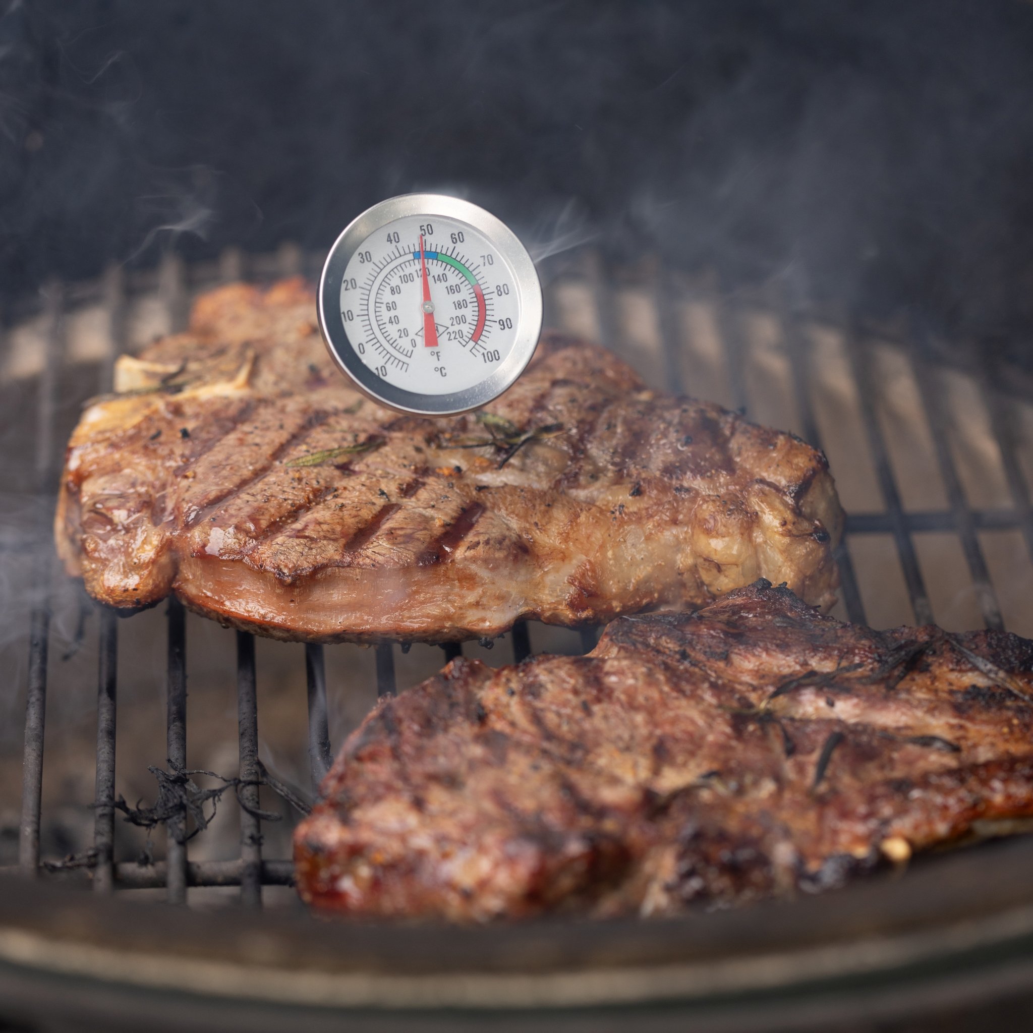 Front view of the thermometer dial above two grilled steaks, with light smoke rising from the grate