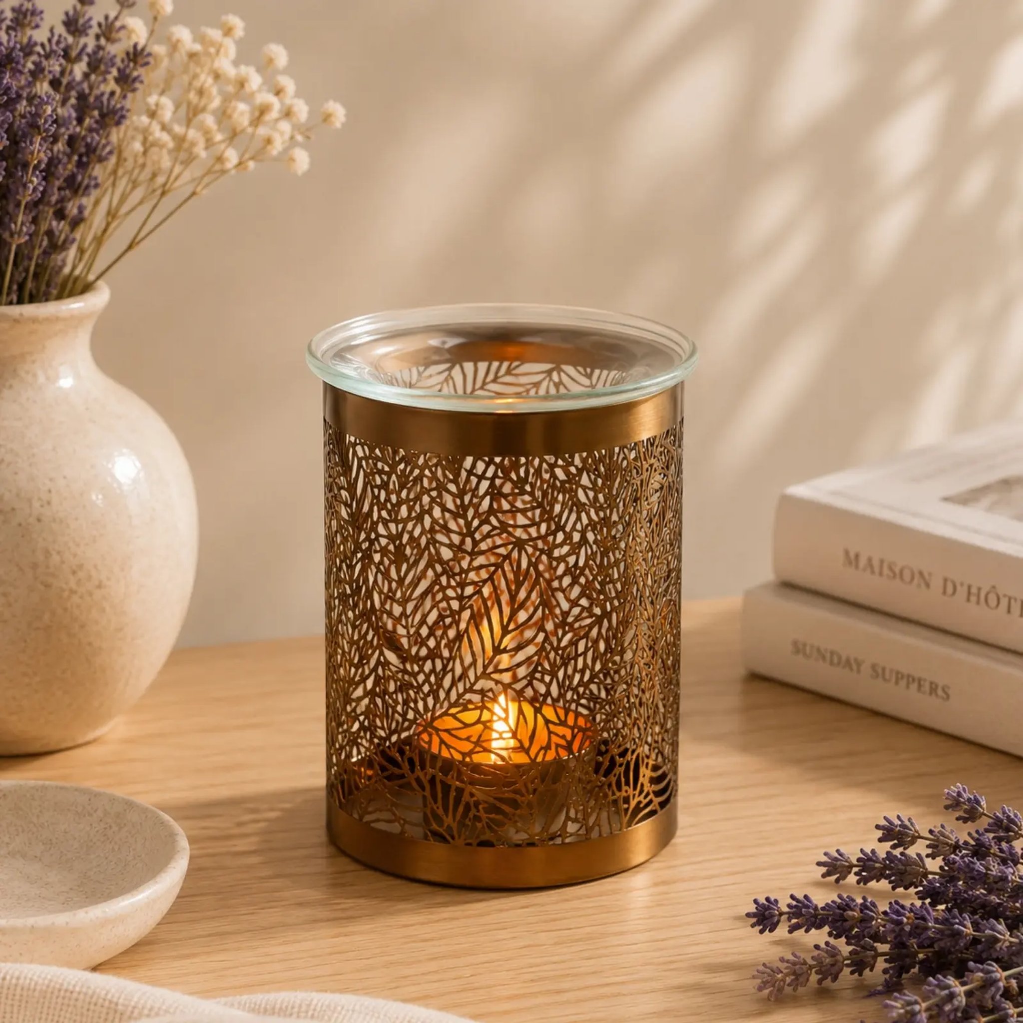 Gold-coloured oil burner with leaf pattern and lit tealight beside lavender and books