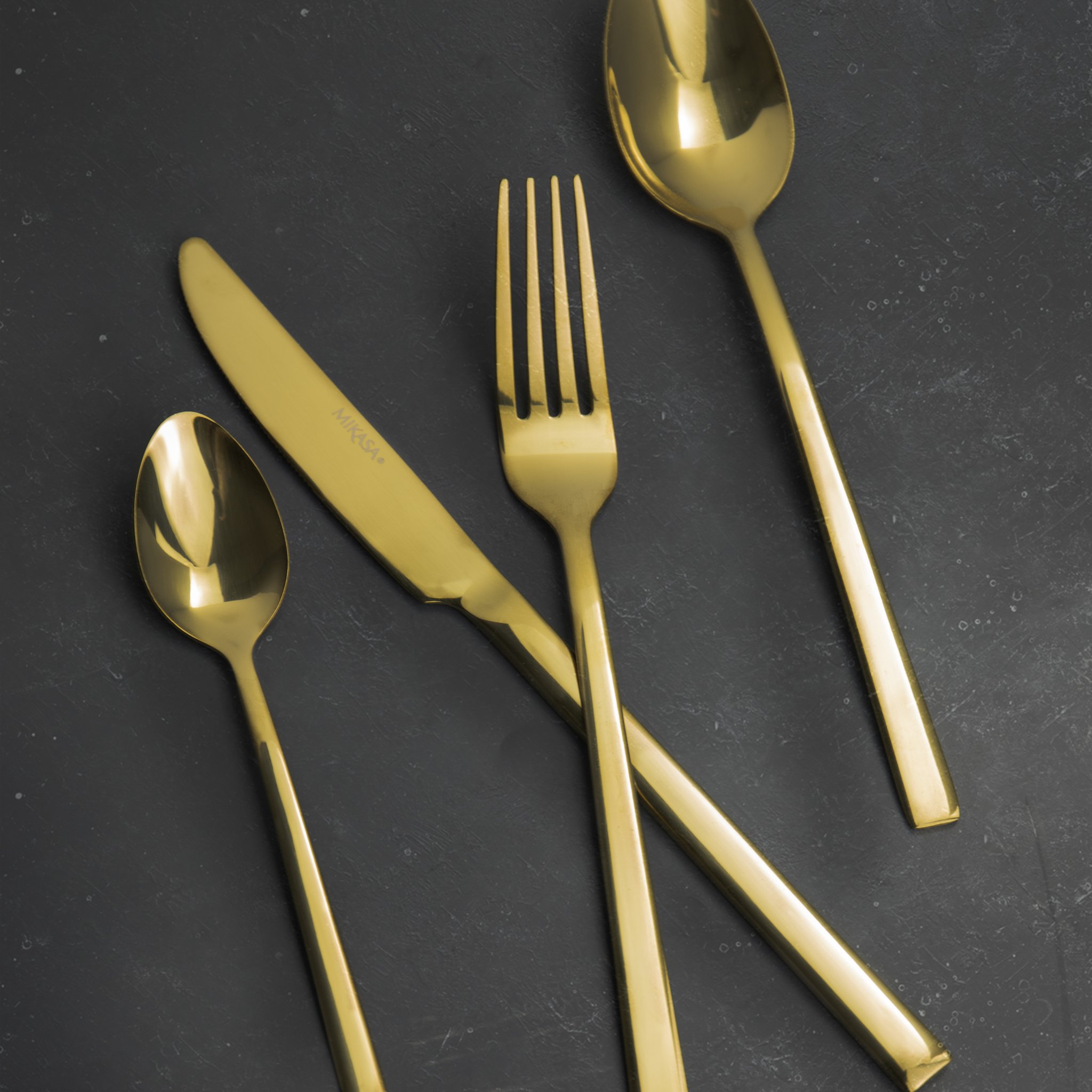 Goldene Löffel, Gabel und Messer übereinander auf dunkler Schieferplatte
