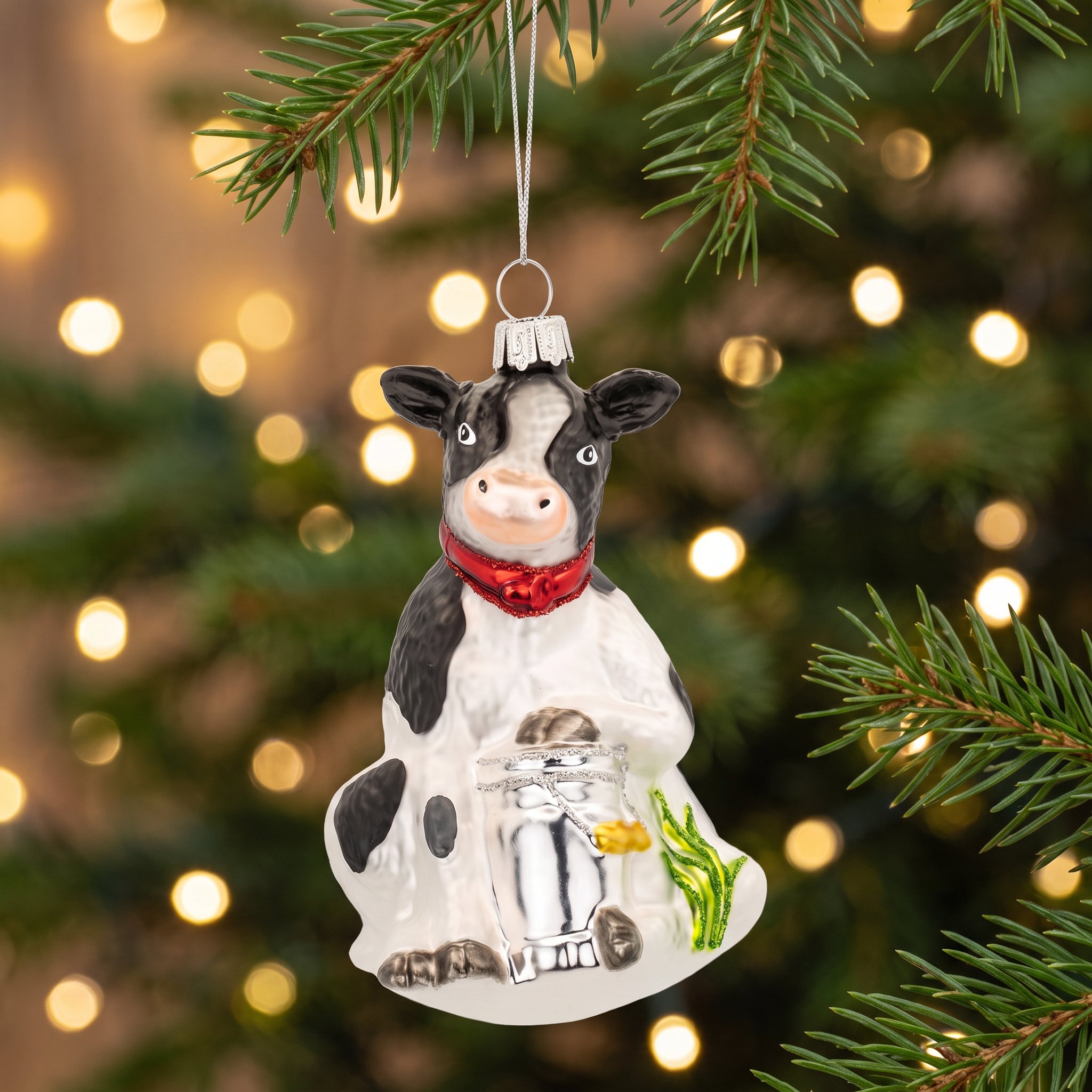 Vache laitière en verre au foulard rouge suspendue à une branche de sapin devant des lumières
