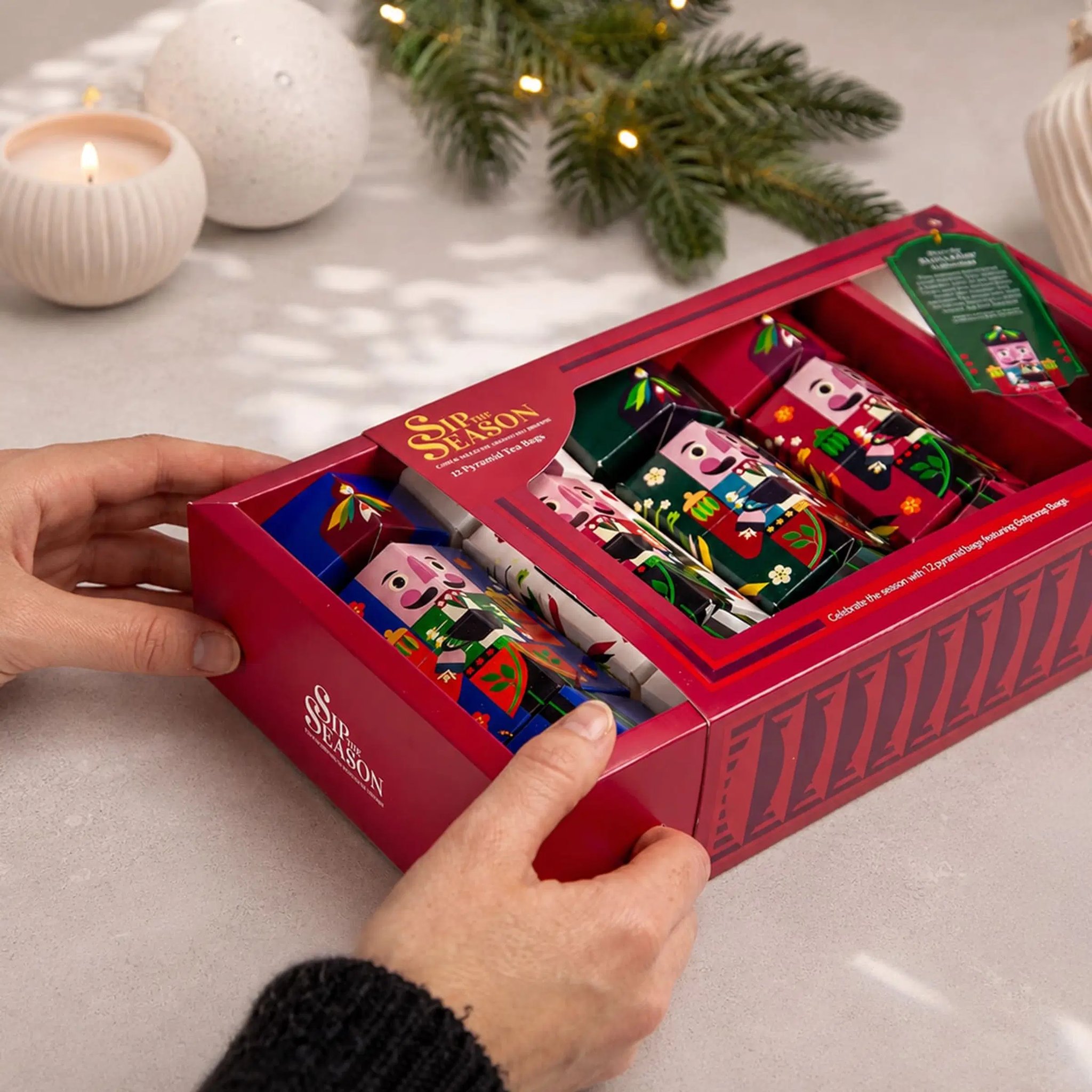 Hands opening the organic Nutcracker Collection tea gift box on a table with candles