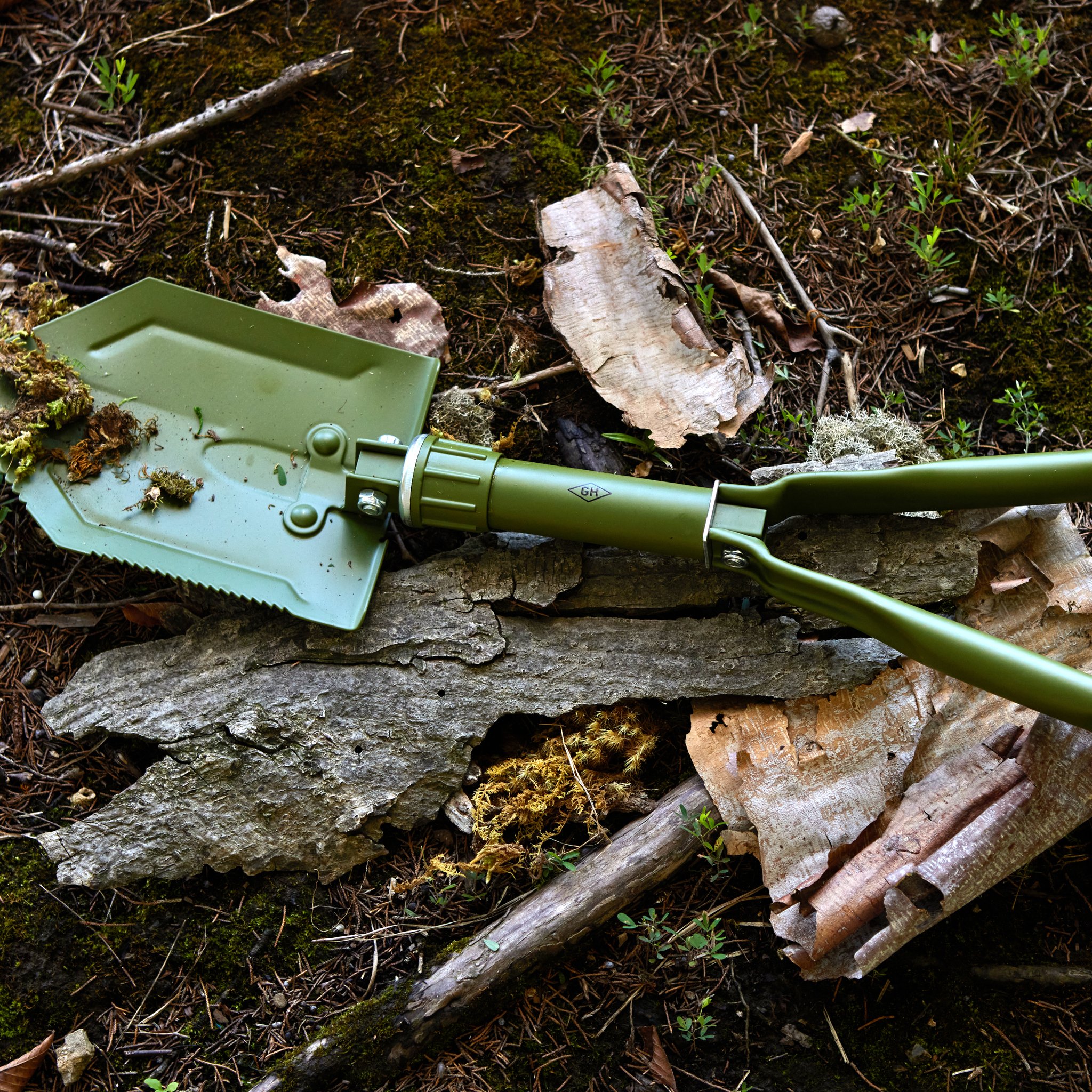 Folding shovel lying on a forest floor among bark, moss and pine needles, with a little soil clinging to the blade