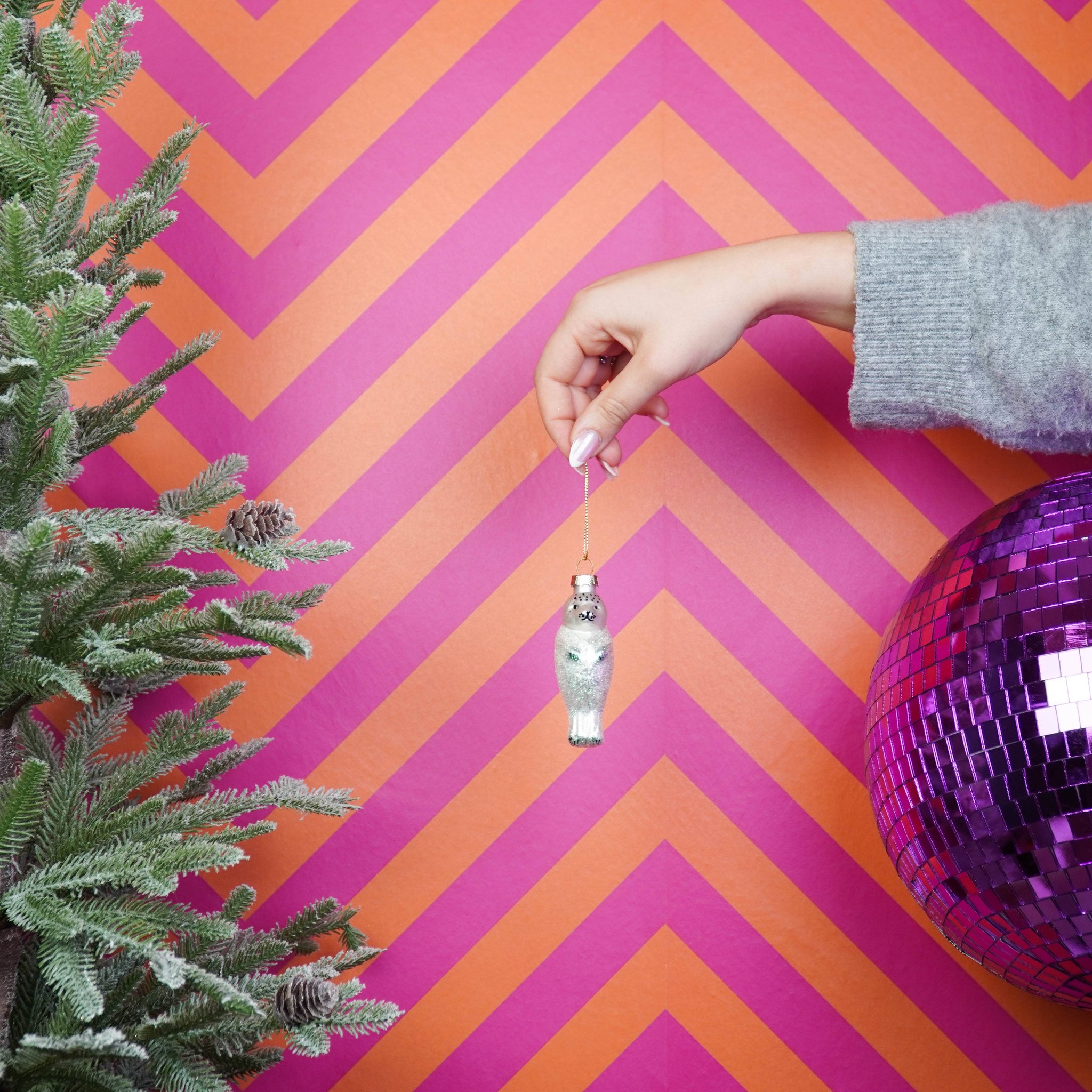 Robbe Weihnachtsanhänger in Silber-Weiss wird vor pinkem Hintergrund mit Discokugel gehalten