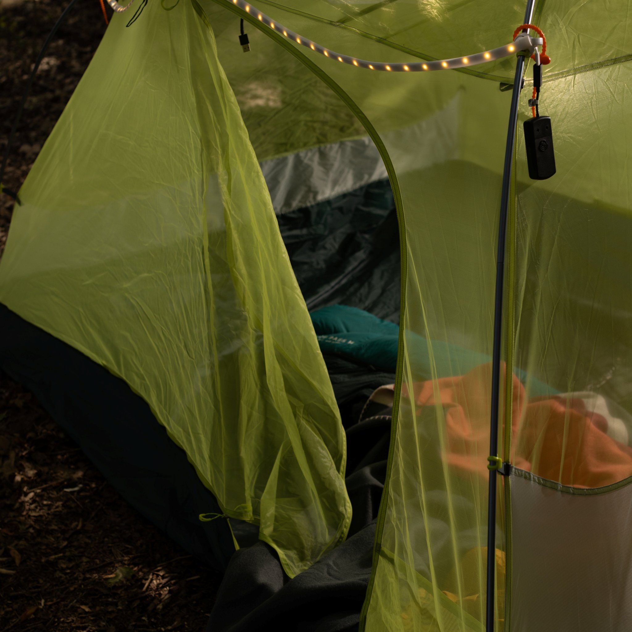 Offener Zelteingang mit Moskitonetz, oben am Gestänge der LED-Lichtschlauch, innen Schlafsack und Ausrüstung