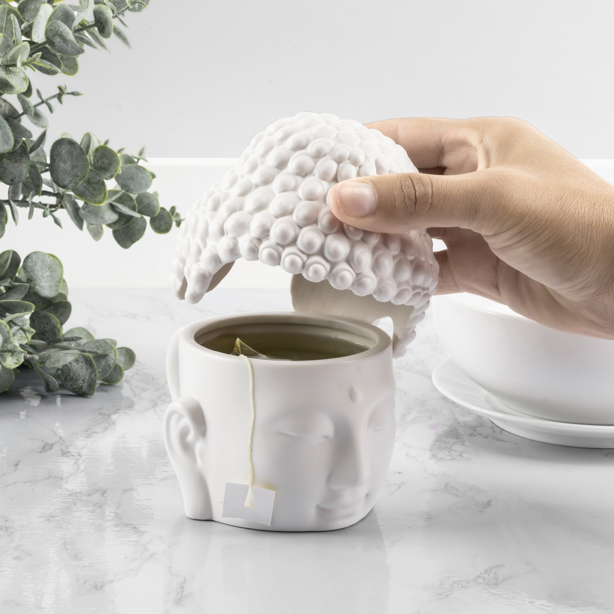 Main soulevant le couvercle du mug Bouddha, sachet de thé qui dépasse